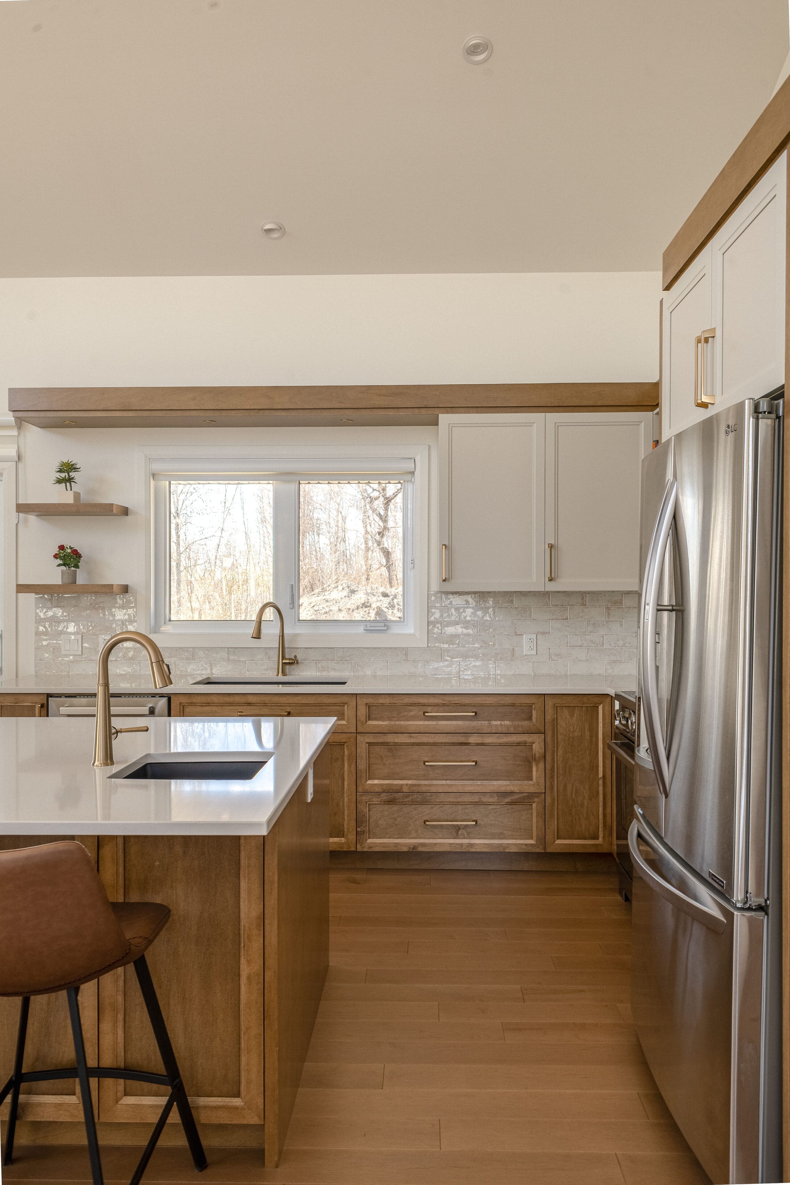 Cuisine moderne avec des planchers en bois clair, des armoires blanches et en bois, un réfrigérateur en acier inoxydable, des luminaires dorés, des comptoirs blancs, une fenêtre au-dessus de l'évier et un tabouret de bar marron sur l'îlot.