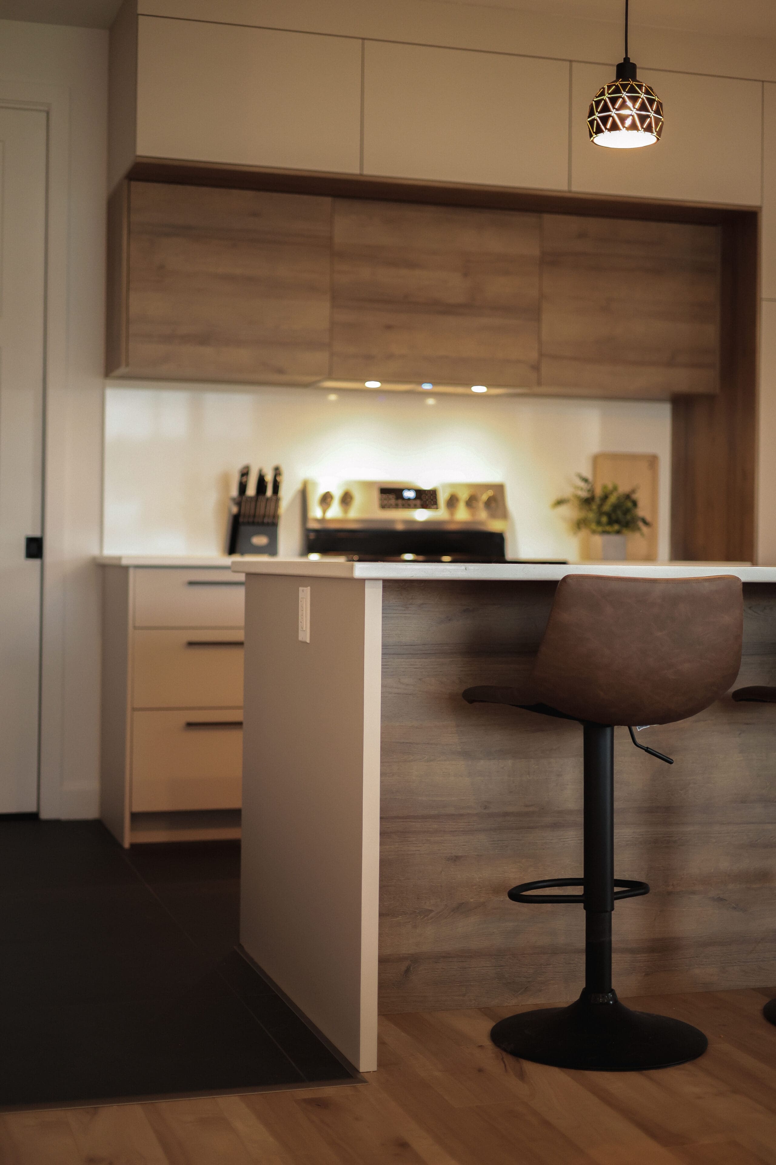Cuisine moderne avec des armoires en bois, un comptoir blanc, une cuisinière avec un bloc de couteaux et une plante à côté, et un tabouret de bar pivotant marron sur l'îlot. Une lampe suspendue est accrochée au-dessus de l'îlot.