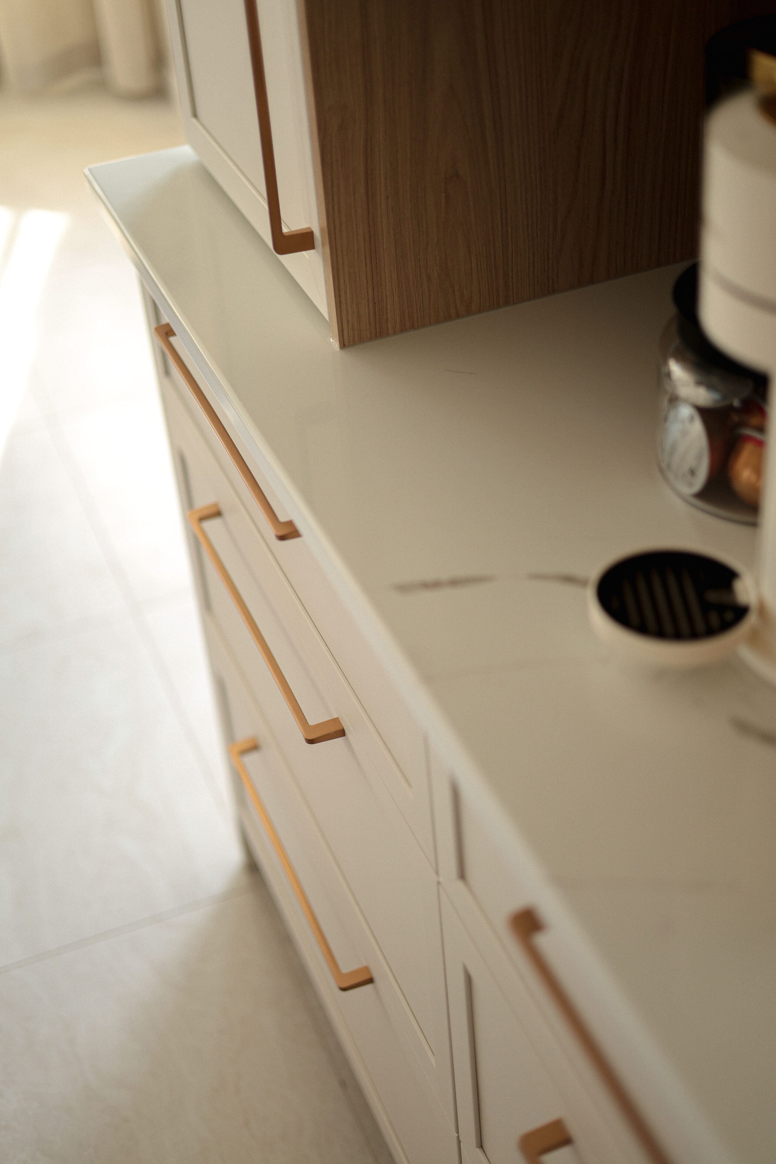 Gros plan sur un comptoir de cuisine moderne avec des meubles blancs et des poignées dorées, avec une surface en marbre et une machine à café avec un pot de capsules de café.