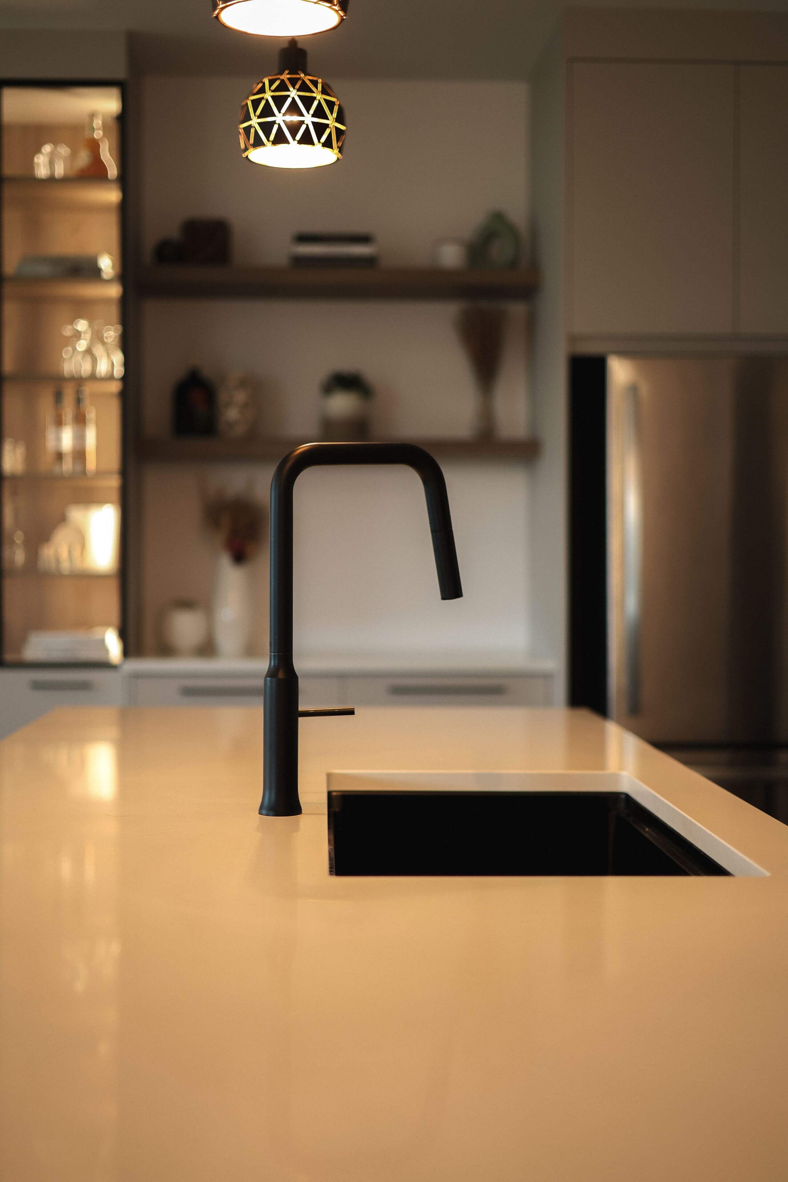 Une cuisine moderne avec un robinet noir élégant sur un îlot de couleur claire, des étagères ouvertes en bois avec de la décoration, des armoires et un réfrigérateur en acier inoxydable en arrière-plan sous un éclairage chaud.