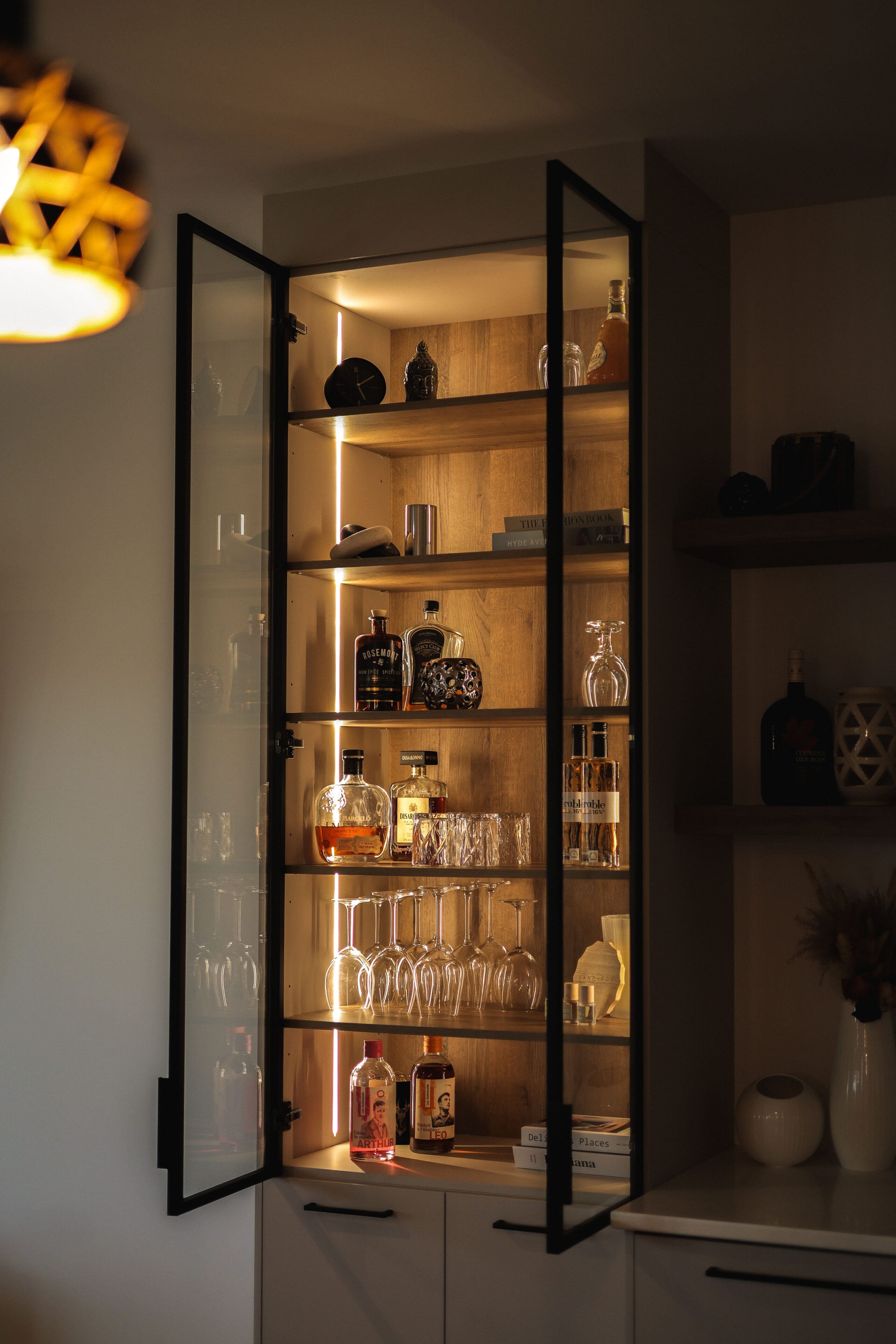 Une armoire vitrée avec des étagères présentant diverses bouteilles d'alcool, de la verrerie et des carafes, le tout éclairé par une lumière intérieure chaude. Certaines bouteilles et certains verres sont bien rangés sur les étagères.