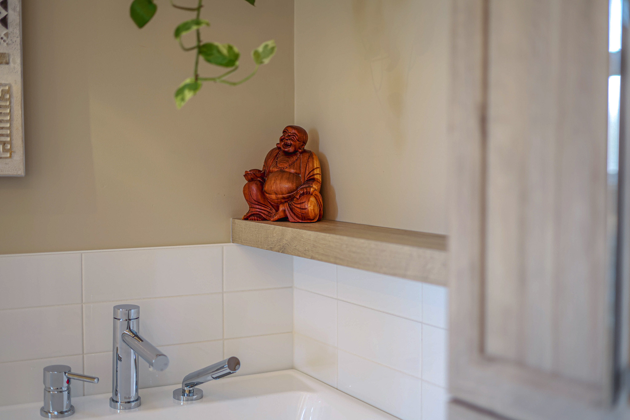 Une petite statue de Bouddha en bois est posée sur une étagère au-dessus d'un lavabo de salle de bains doté d'un robinet moderne. Une plante à feuilles est suspendue, partiellement visible du dessus. Le mur est beige et des carreaux blancs bordent l'arrière du lavabo.