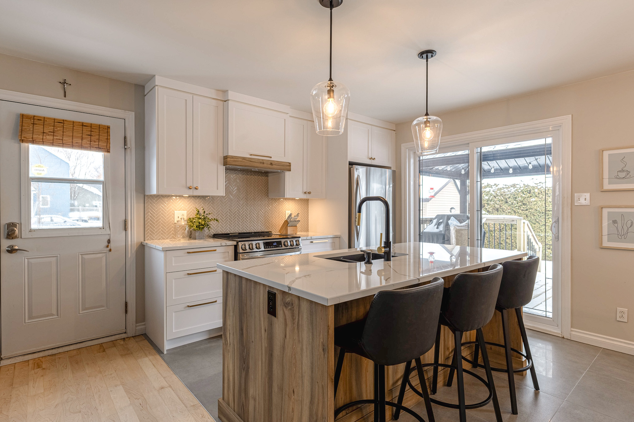 Cuisine moderne avec des armoires blanches, un grand îlot avec des accents en bois, trois tabourets de bar noirs, des lampes suspendues, des appareils électroménagers en acier inoxydable et des portes coulissantes en verre menant à un patio extérieur.