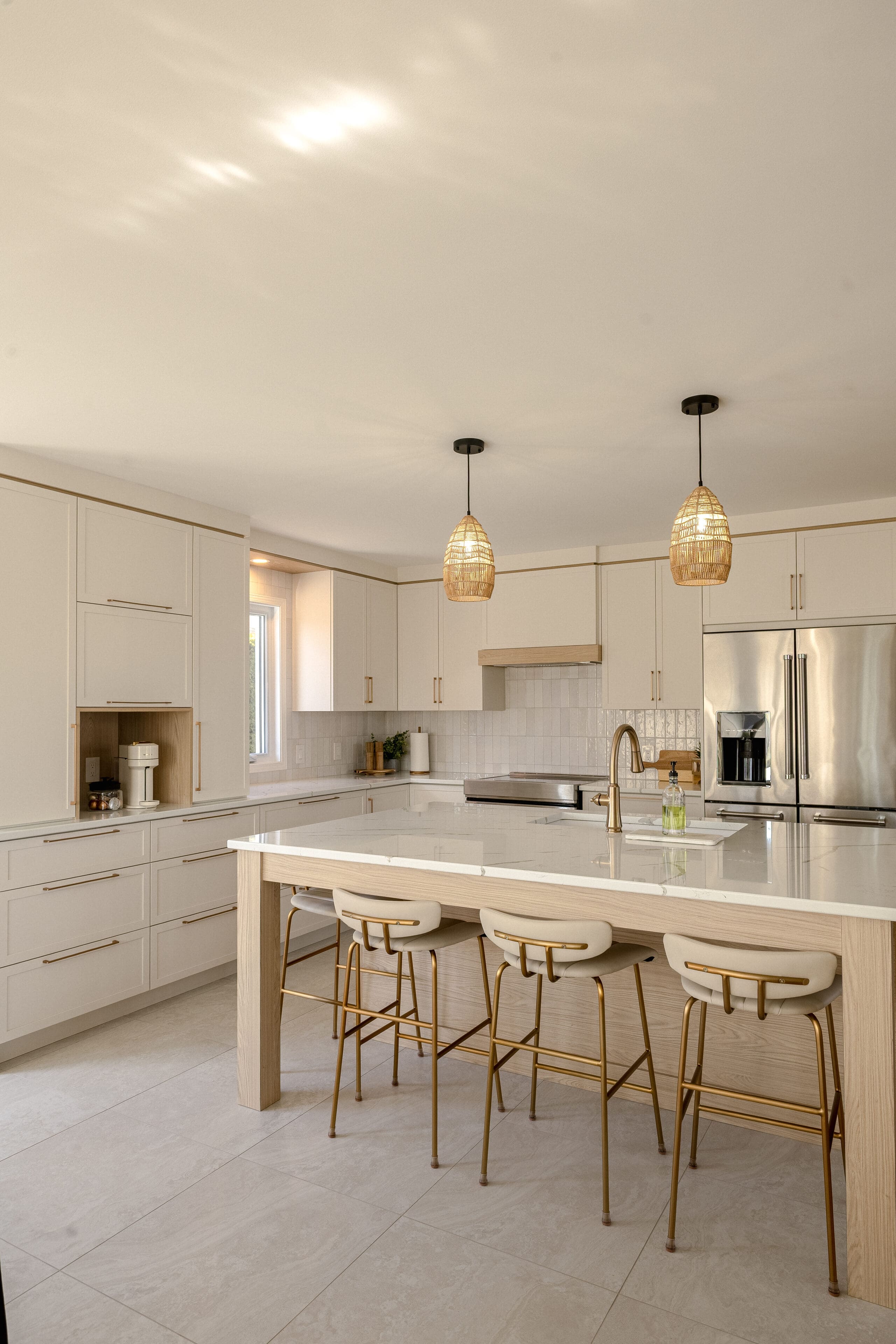 Cuisine moderne avec des armoires en bois clair, un grand îlot avec un comptoir blanc, trois tabourets de bar rembourrés, des appareils électroménagers en acier inoxydable et deux lampes suspendues tressées suspendues au-dessus de l'îlot. La lumière naturelle remplit l'espace.