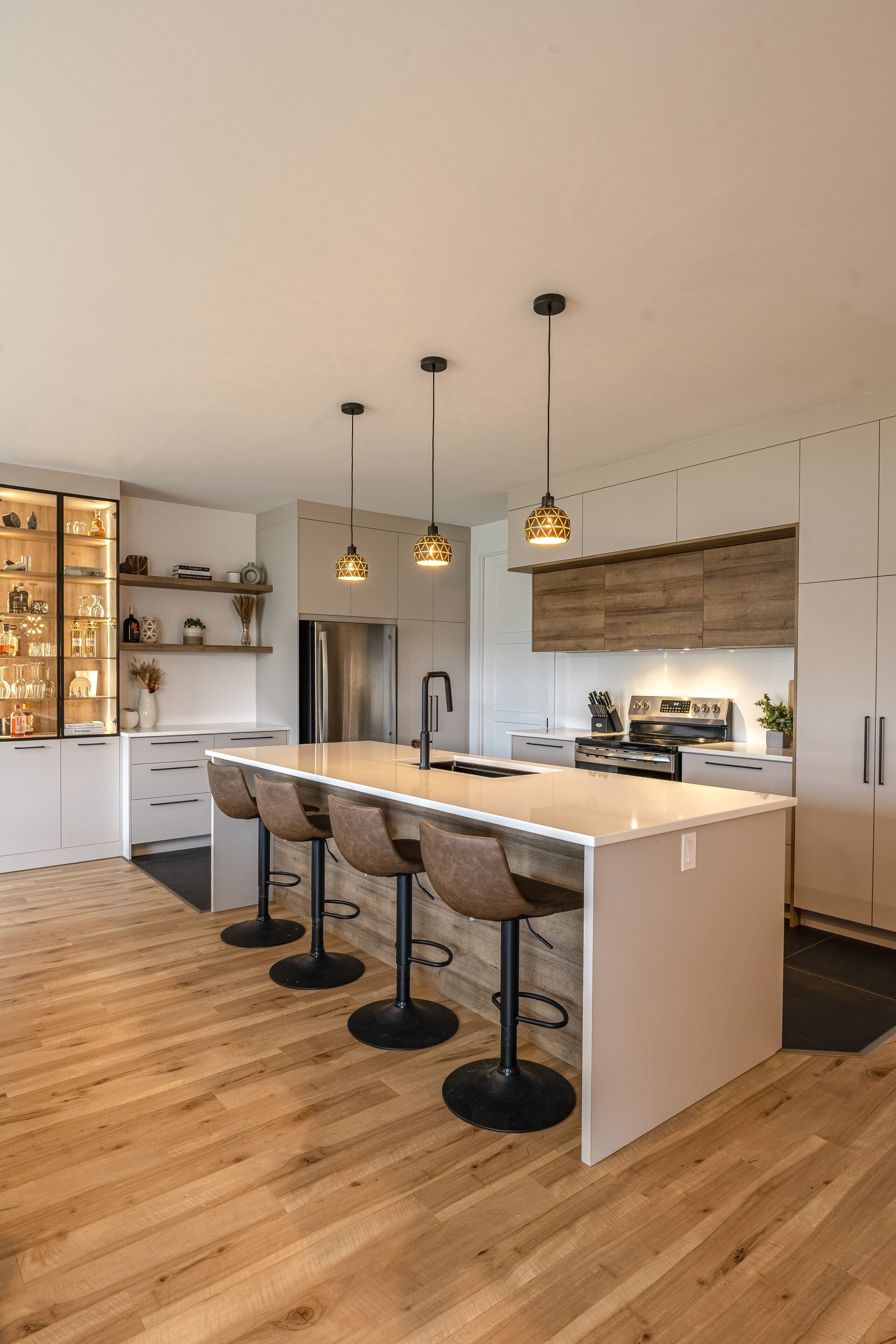 Cuisine moderne avec des planchers en bois clair, un grand îlot blanc avec quatre tabourets de bar bruns, des lampes suspendues au-dessus, des appareils encastrés, des armoires légères et des étagères ouvertes présentant de la vaisselle et de la décoration.