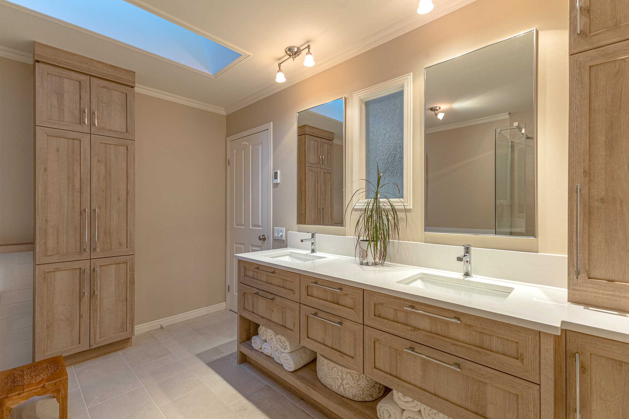 Salle de bains moderne avec double lavabo, armoires en bois, miroirs, puits de lumière et murs de couleur neutre. Une plante en pot est posée sur le plan de travail et les serviettes sont soigneusement rangées sur des étagères sous les lavabos.