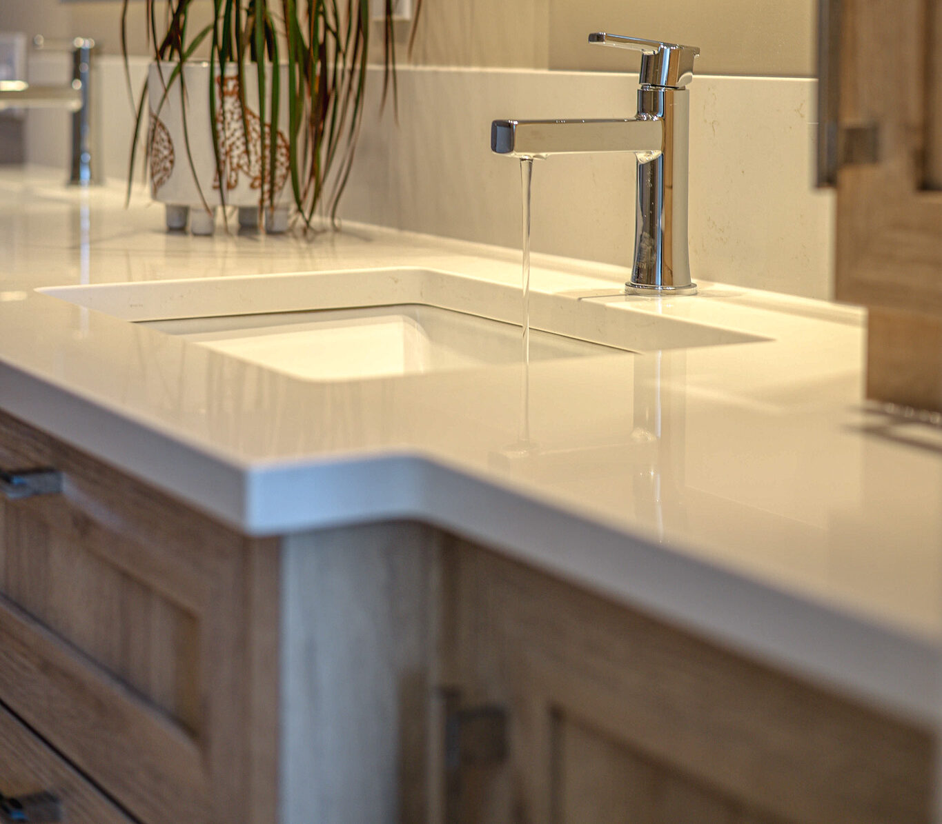 Un lavabo de salle de bain moderne avec un robinet chromé élégant laissant couler l'eau, posé sur un comptoir de quartz blanc. Une grande plante feuillue et un meuble-lavabo en bois sont visibles, avec un miroir et des éléments décoratifs en arrière-plan.