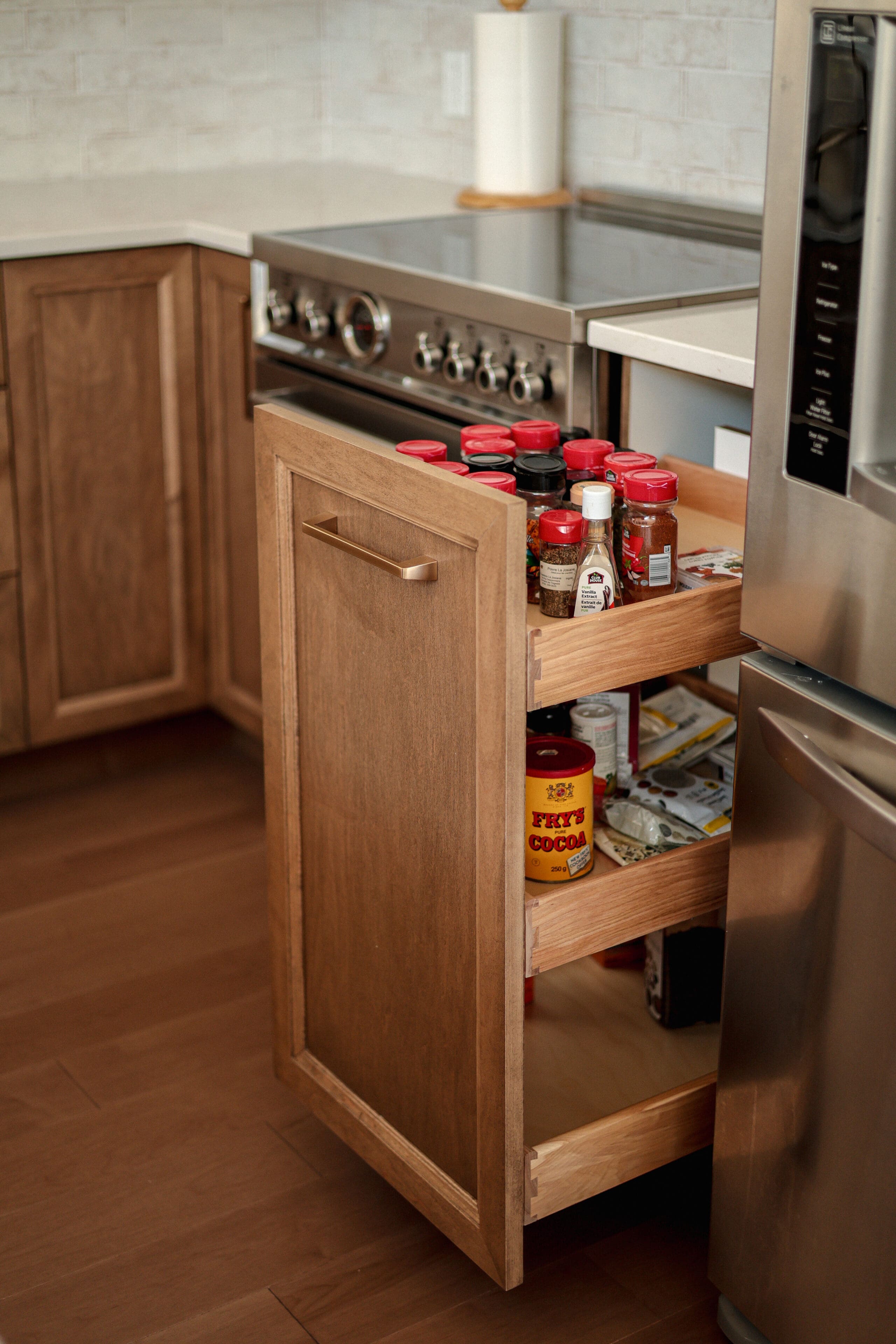 Dans une cuisine, un tiroir de garde-manger en bois est ouvert, révélant des étagères remplies de pots d'épices, de sauces et de conserves à côté d'une cuisinière et d'un réfrigérateur.