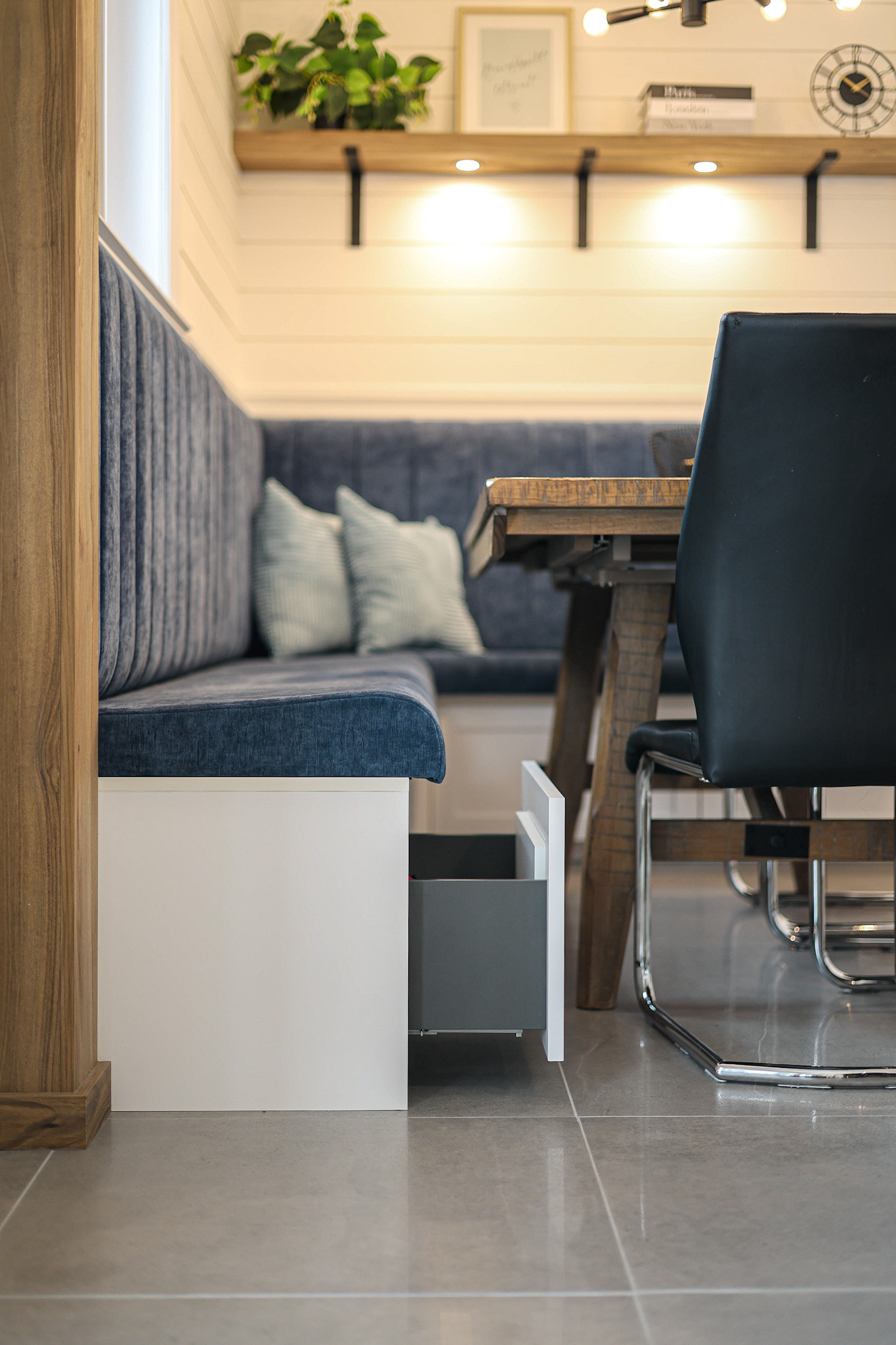 Un coin repas moderne avec un banc rembourré bleu encastré doté d'un tiroir de rangement extractible, une table en bois, une chaise noire, des coussins et des étagères murales avec de la décoration et une plante.
