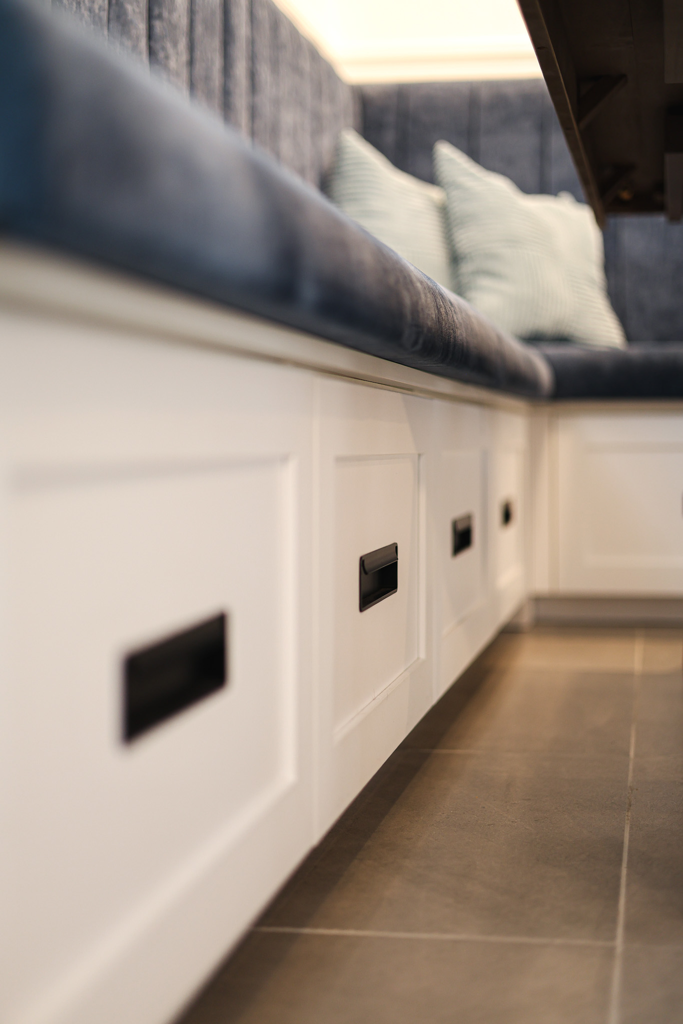 Banc de rangement blanc encastré avec des poignées noires, surmonté d'une assise coussinée bleue et de coussins rayés, le tout posé sur un sol carrelé. L'accent est mis sur les tiroirs du banc et sur son design moderne et confortable.