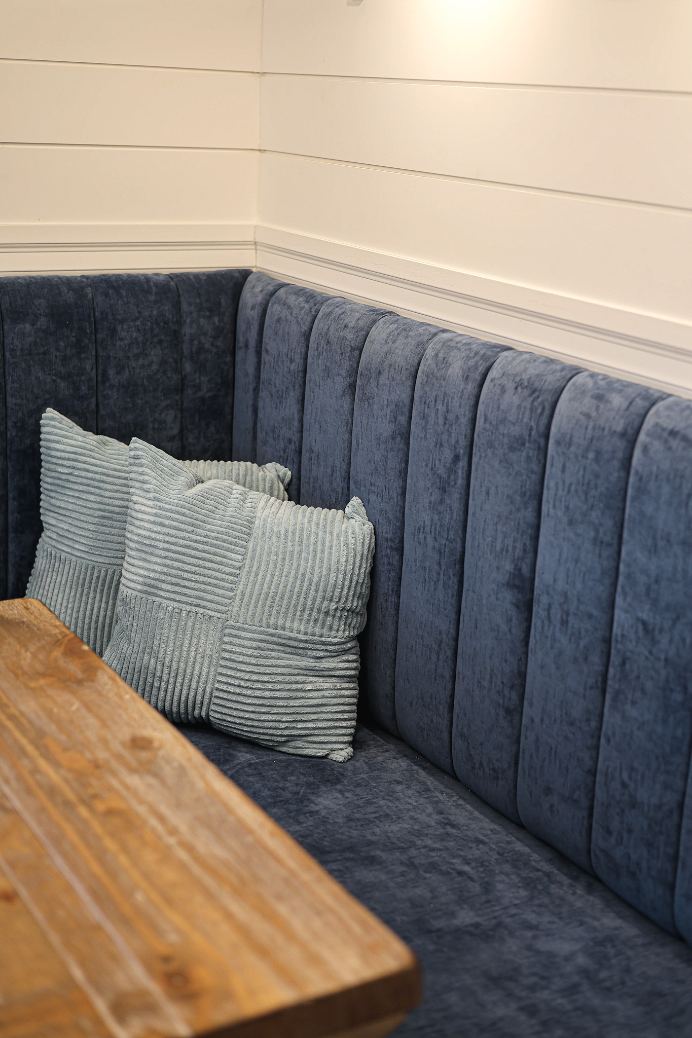 Une cabine d'angle confortable avec un revêtement bleu foncé à rayures verticales, deux coussins en velours côtelé bleu clair et une table en bois, le tout posé sur des murs blancs en shiplap.