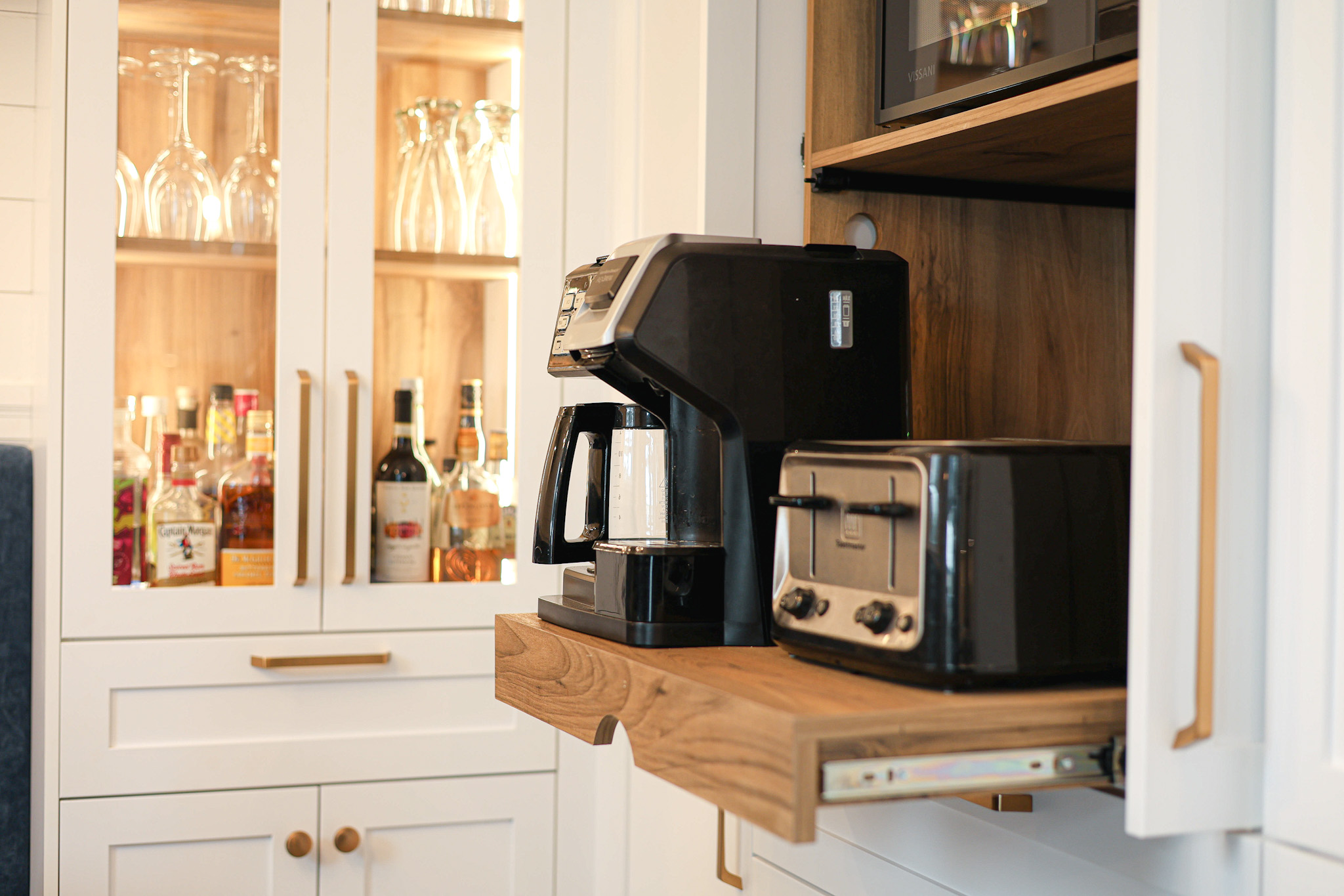 La cuisine est équipée d'une étagère coulissante en bois avec une cafetière et un grille-pain. Derrière, des armoires blanches avec des portes en verre présentent des verres et diverses bouteilles. La scène est chaleureusement éclairée et organisée.