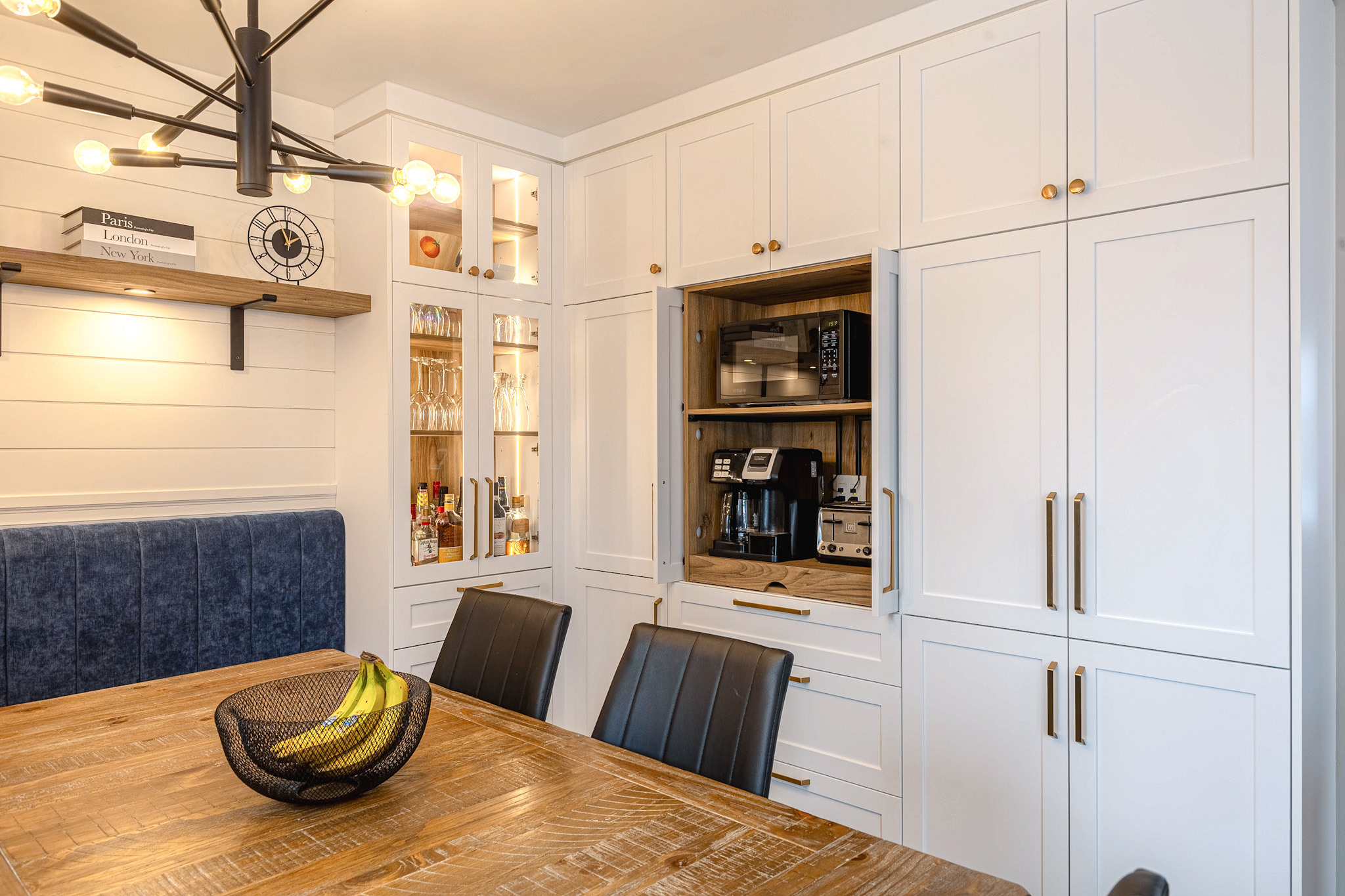 Cuisine moderne avec des armoires blanches, une cafetière encastrée, une table à manger en bois avec un bol de bananes, des chaises noires, une banquette bleue coussinée et un lustre contemporain noir avec des ampoules apparentes.