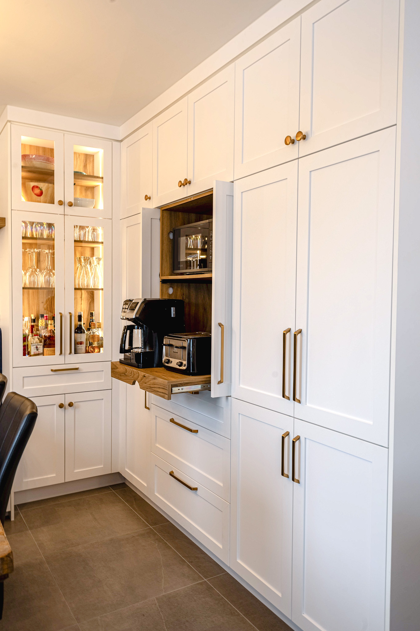 Une cuisine moderne avec des armoires blanches, des poignées dorées et une étagère coulissante dissimulée contenant une machine à café et un grille-pain ; des verres et des bouteilles sont exposés dans une armoire éclairée sur la gauche.