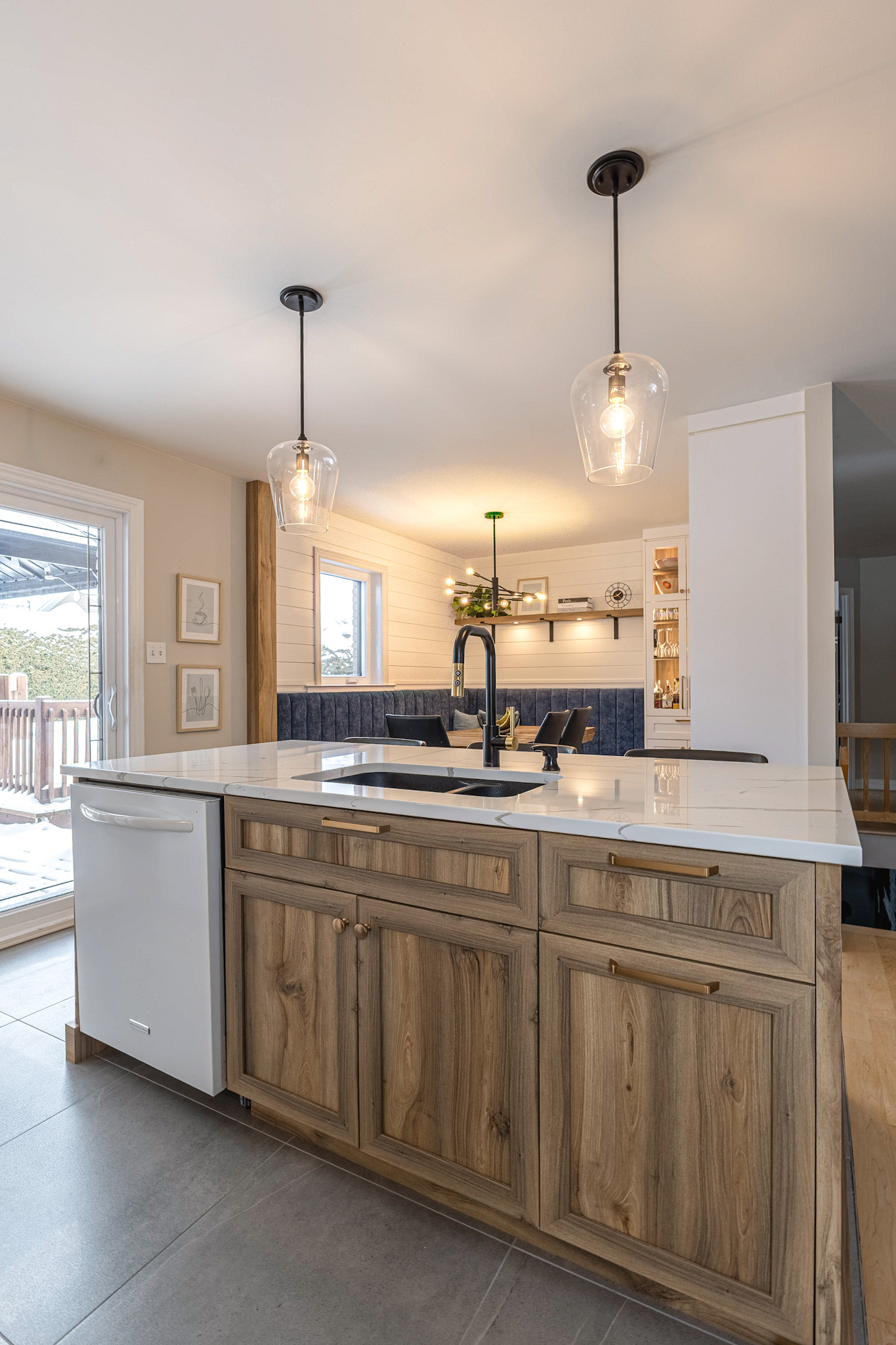 Une cuisine moderne avec un îlot en bois, un comptoir blanc, un évier de ferme, un robinet noir et deux lampes suspendues. À l'arrière-plan, un coin repas avec une table, une banquette intégrée et des étagères.
