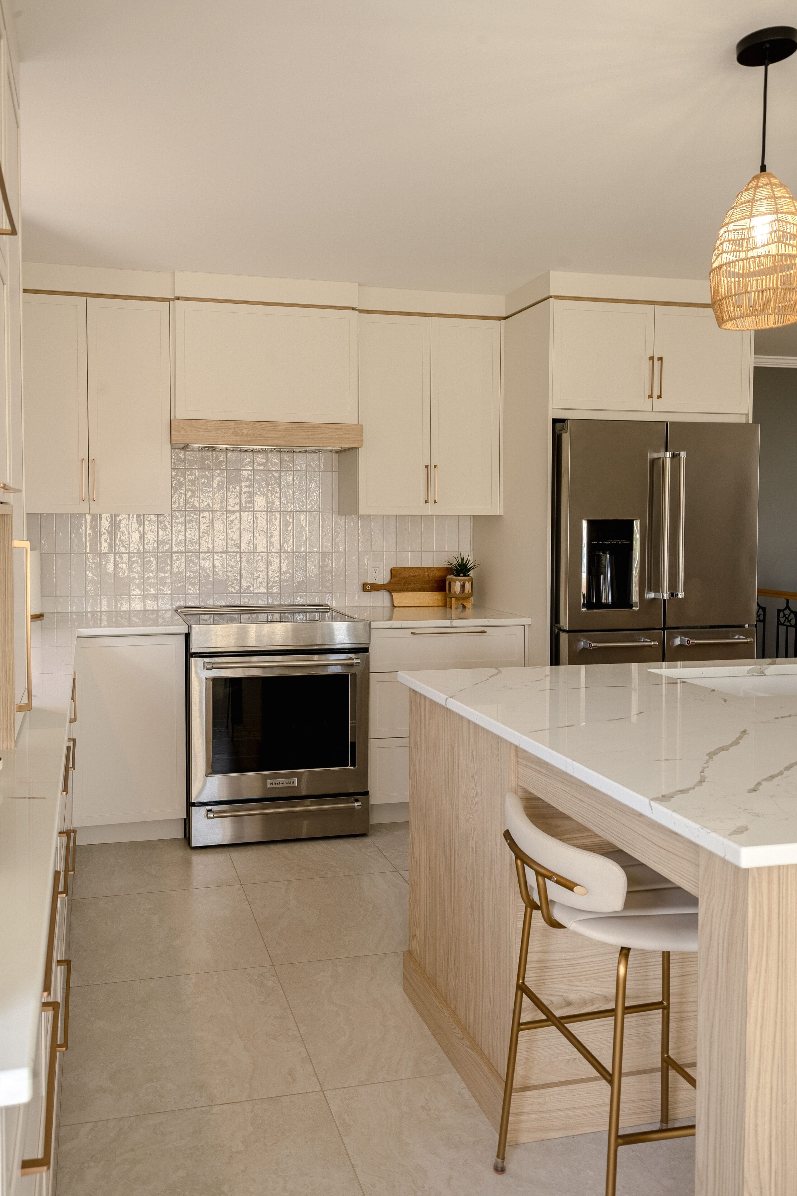 Cuisine moderne avec des armoires en bois clair et blanc, des appareils électroménagers en acier inoxydable, un îlot en marbre avec une chaise blanche, un dosseret en carrelage et une lampe suspendue en osier accrochée au plafond.