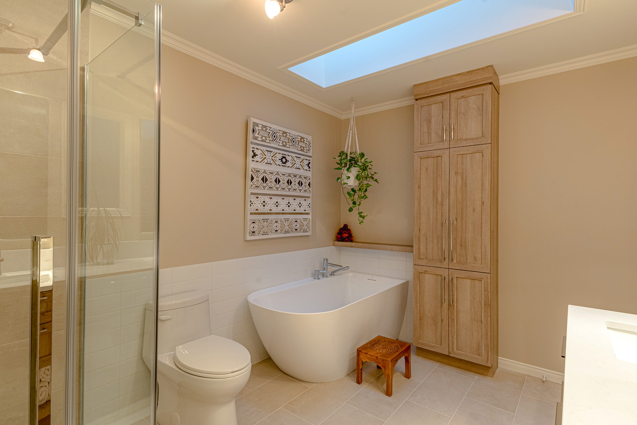 Salle de bains moderne avec une baignoire blanche autoportante, des toilettes, une douche en verre, un grand meuble en bois, des œuvres d'art murales, une plante suspendue, un petit tabouret en bois et un puits de lumière au-dessus qui laisse entrer la lumière naturelle.
