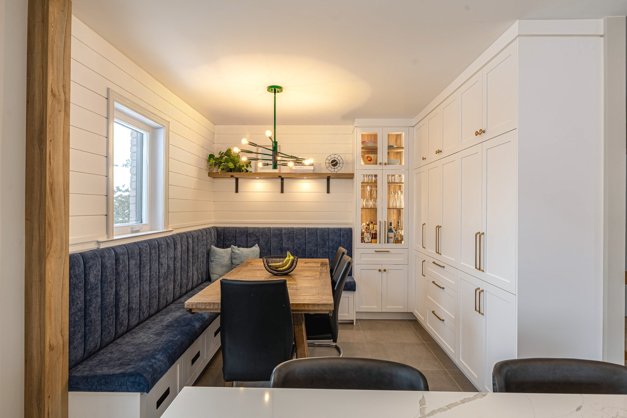Un coin repas confortable avec une table en bois, une banquette tapissée bleu marine, des chaises noires et un meuble blanc encastré. Un lustre moderne est suspendu au-dessus, et une fenêtre apporte la lumière naturelle dans l'espace.