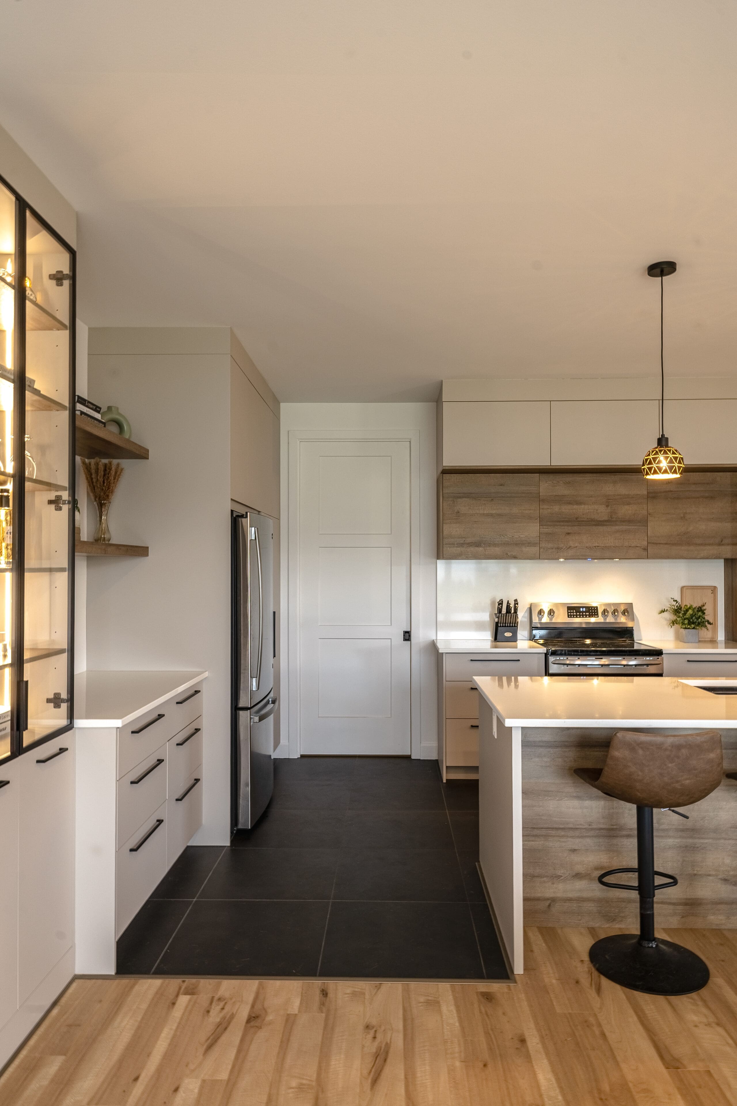 Cuisine moderne avec des armoires en bois et en blanc, des appareils en acier inoxydable, du carrelage noir, un îlot central avec une chaise pivotante marron et une lampe suspendue au-dessus de l'îlot. Des étagères avec des portes en verre se trouvent sur la gauche.