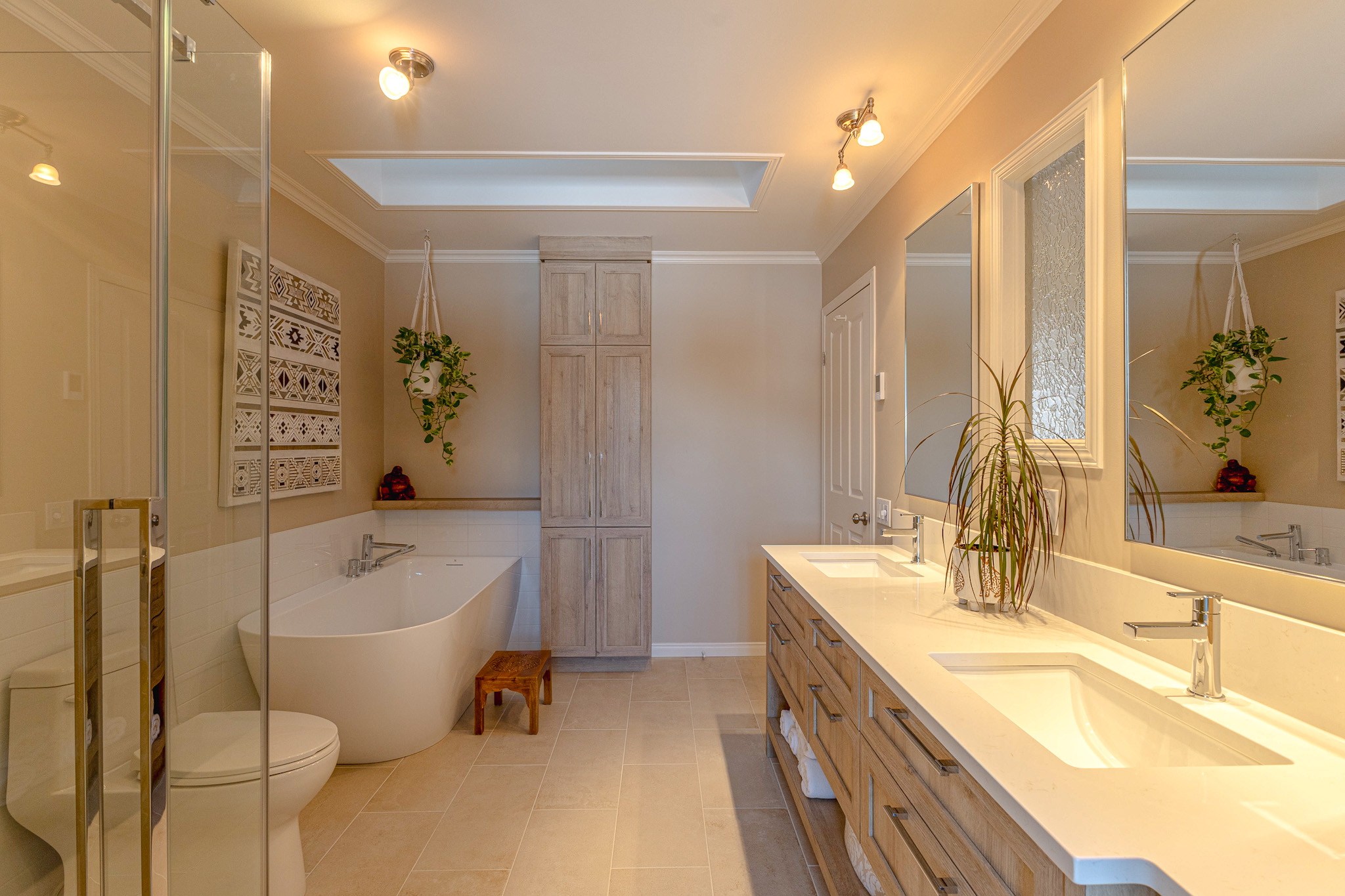 Salle de bains moderne avec double lavabo, grands miroirs, baignoire autoportante, plantes suspendues, armoires en bois, douche en verre et carrelage beige sous un éclairage chaleureux.