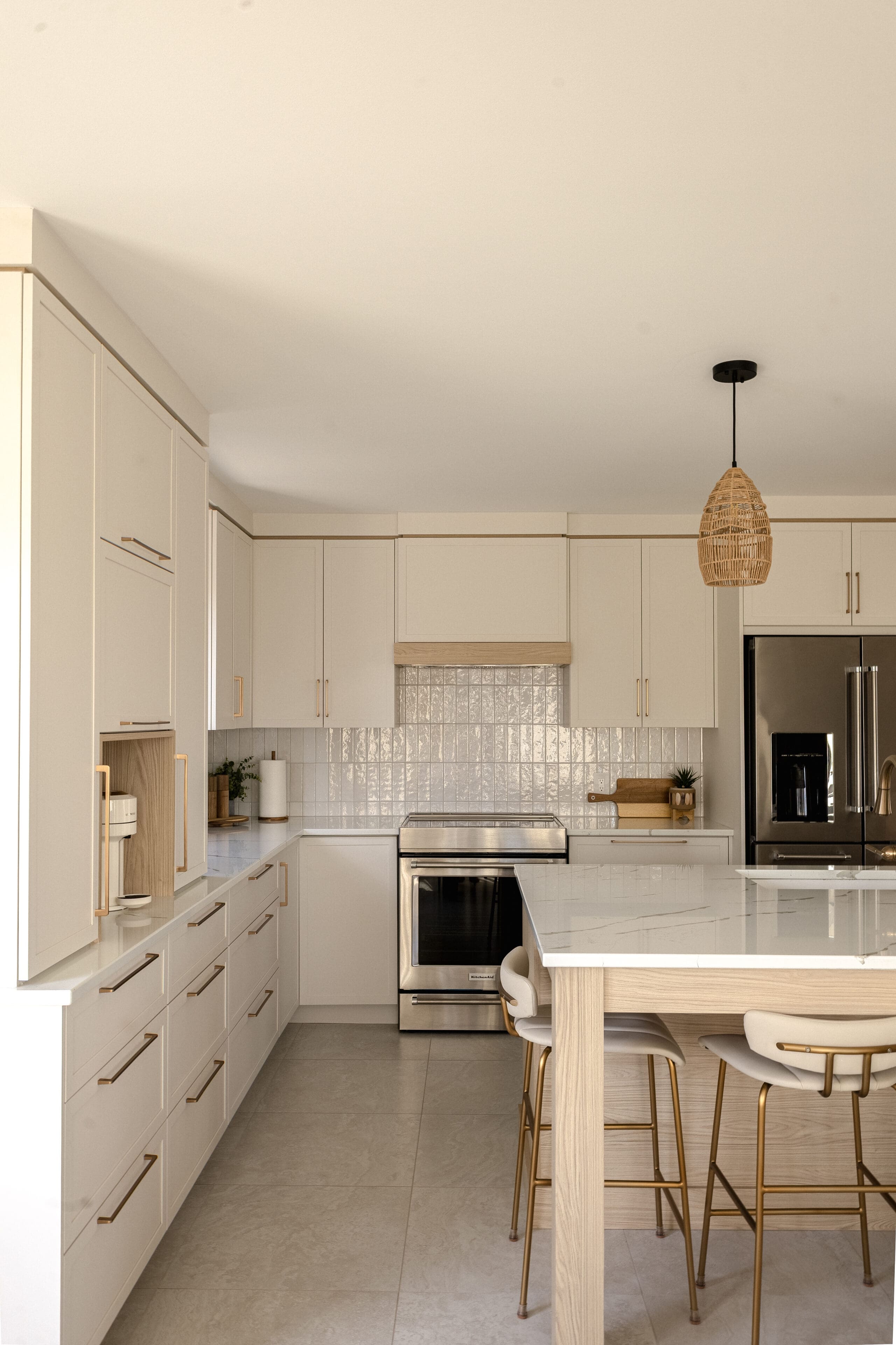 Cuisine moderne avec des armoires blanches, des poignées dorées, des appareils en acier inoxydable, un dosseret carrelé, un grand îlot avec un comptoir blanc et des accents en bois, trois tabourets de bar blancs et une lampe suspendue tissée.