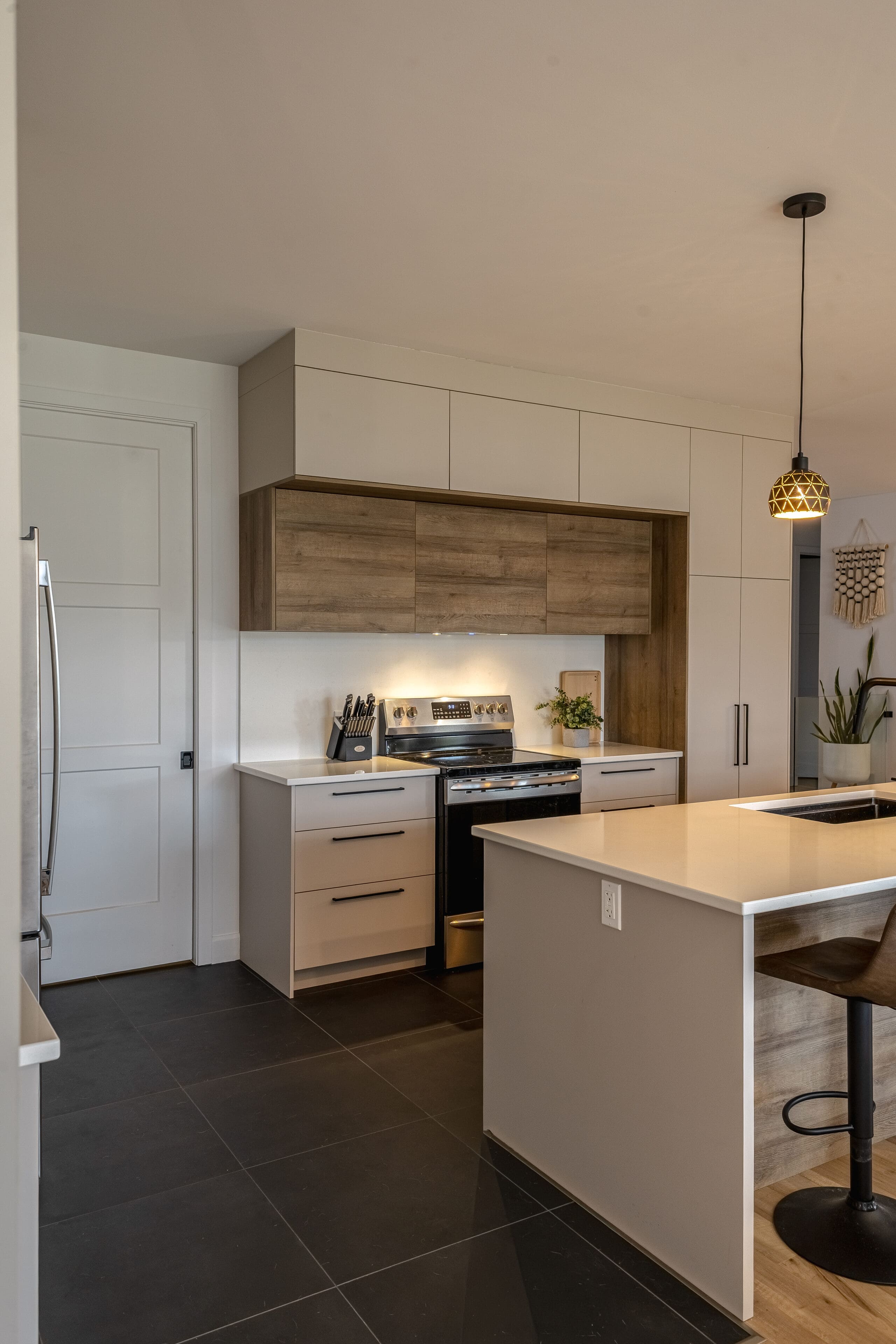 Cuisine moderne avec des armoires blanches et en bois, un four en acier inoxydable et un sol en carrelage noir. Un îlot avec des tabourets de bar est placé sous une lampe suspendue décorative. Une plante et des ustensiles sont posés sur le comptoir.