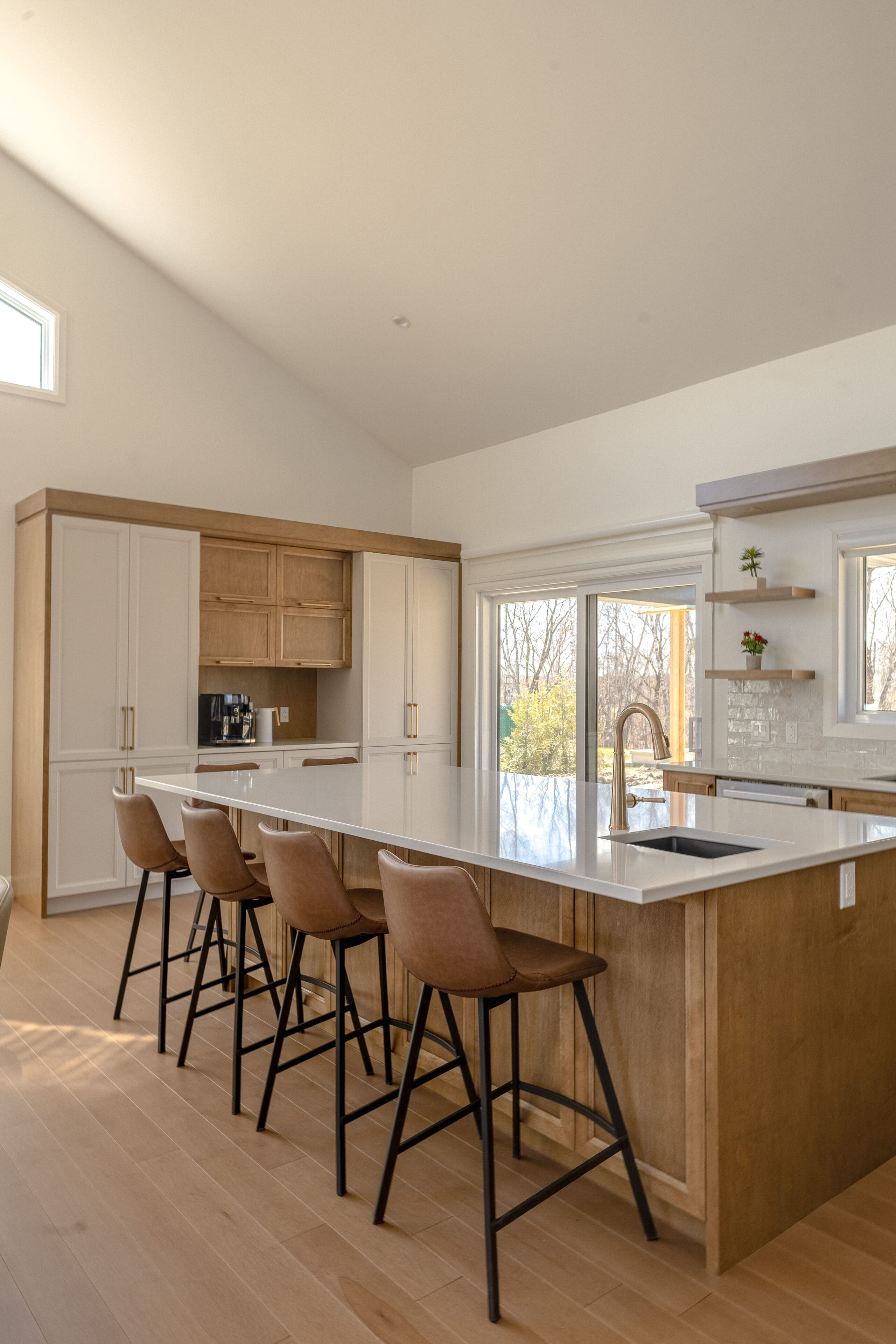 Cuisine moderne avec un sol en bois clair, un grand îlot avec un comptoir blanc, quatre tabourets de bar bruns, des armoires encastrées, une cafetière et de grandes fenêtres laissant entrer la lumière naturelle.
