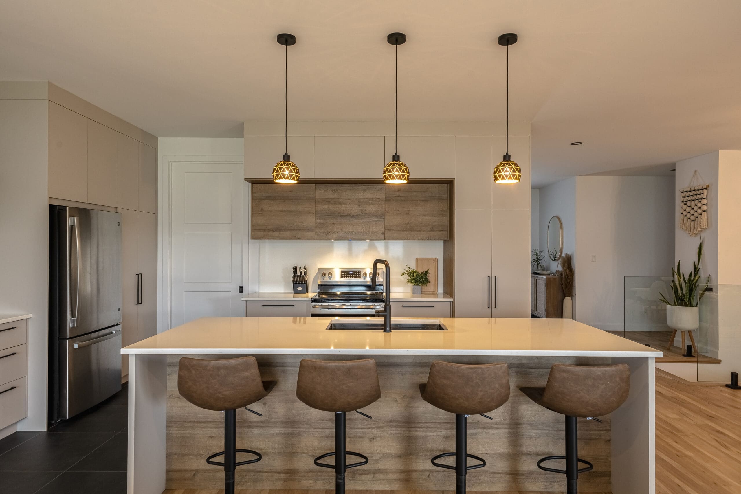 Cuisine moderne avec un îlot central, quatre tabourets de bar marron, des lampes suspendues, des appareils en acier inoxydable, un sol en bois clair et un décor minimaliste dans des tons neutres.