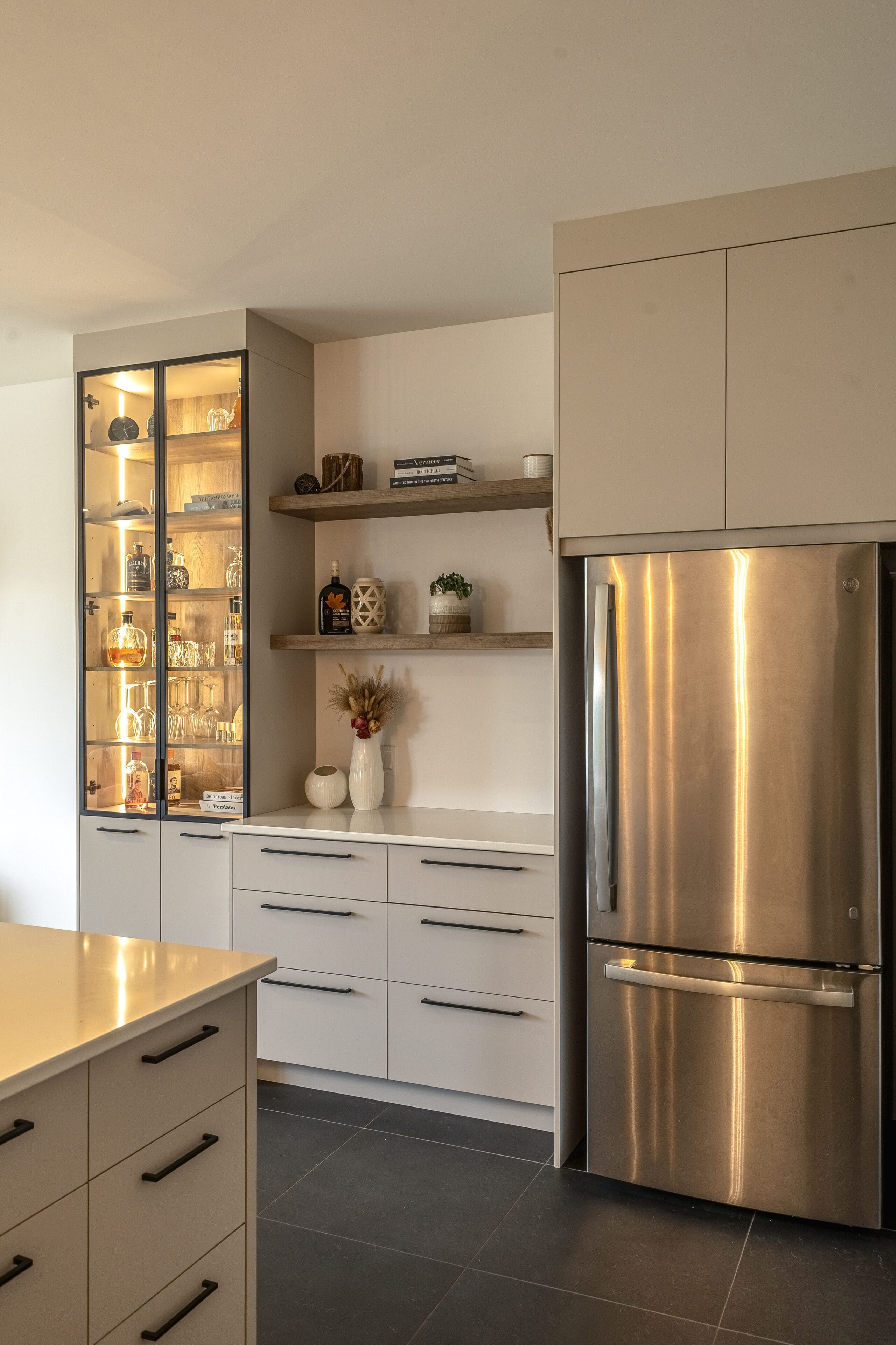 Cuisine moderne avec des armoires claires, un réfrigérateur en acier inoxydable, un meuble bar à façade vitrée avec des étagères éclairées contenant des bouteilles et des verres, des étagères ouvertes en bois avec des objets décoratifs, et un sol en carrelage foncé.