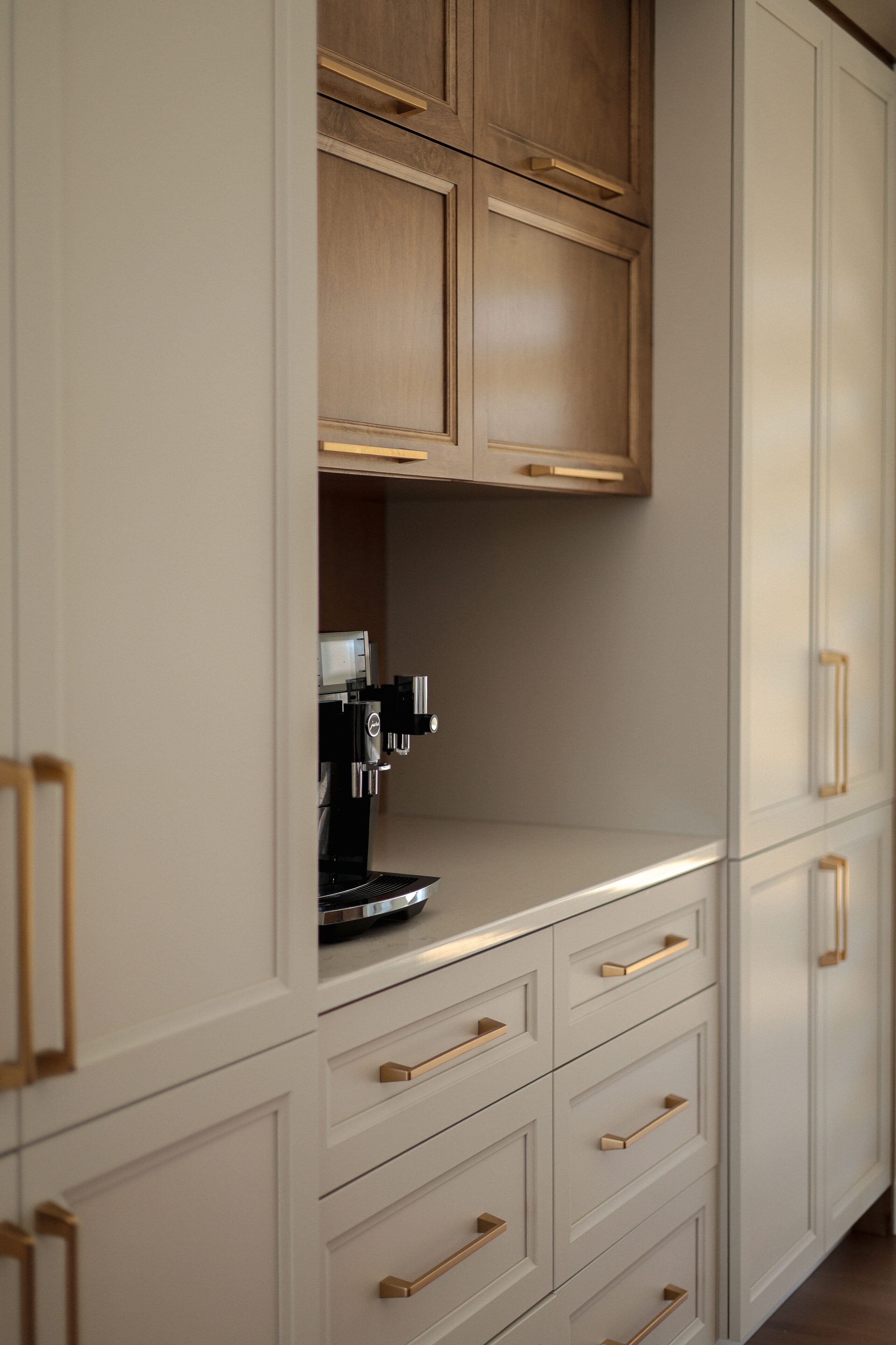 Machine à café encastrée dans un coin cuisine moderne avec des armoires beige clair, des poignées en laiton et des armoires supérieures en bois. L'espace présente des lignes épurées et un design minimaliste.