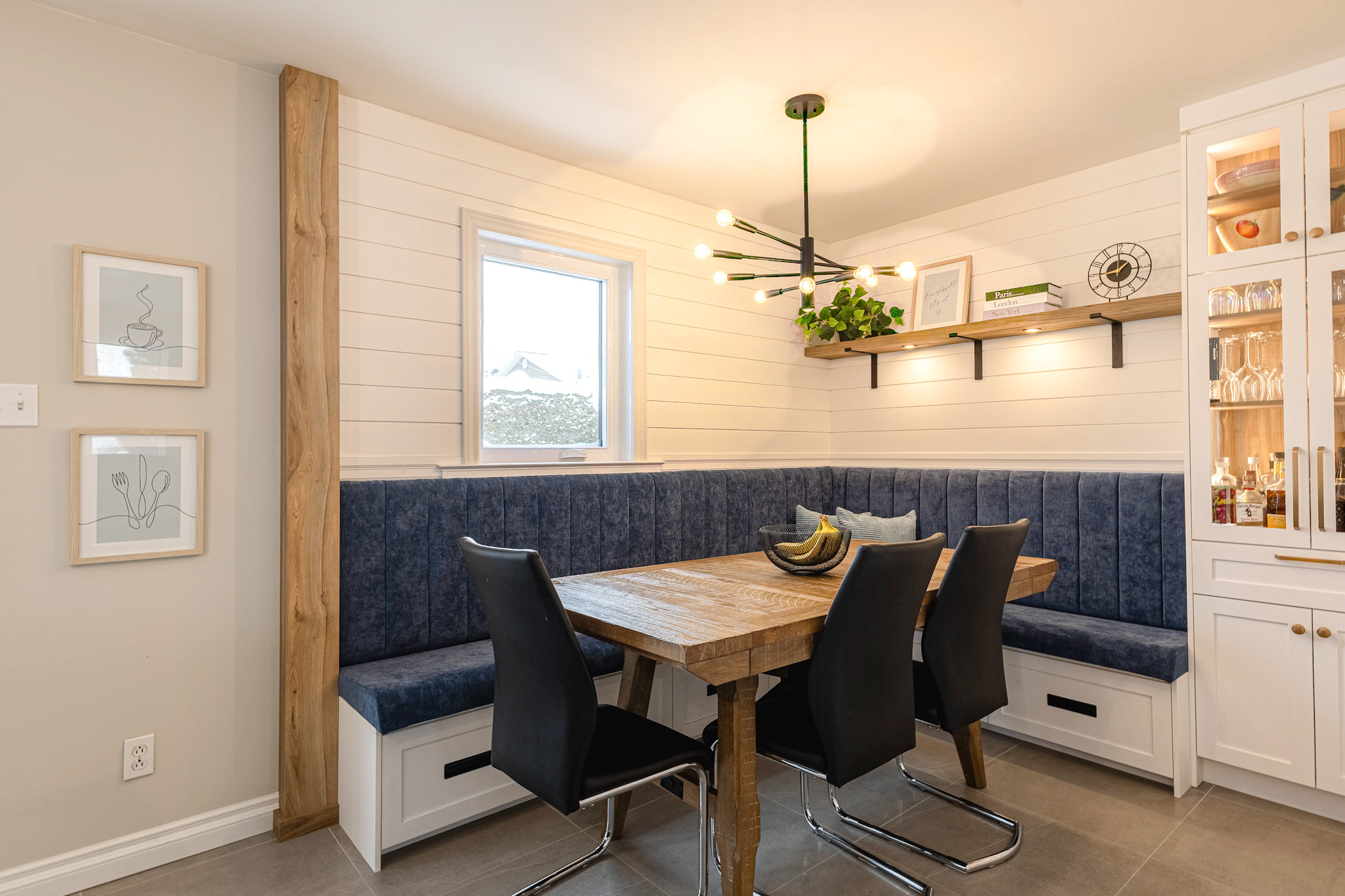 Un coin repas confortable avec une table en bois, quatre chaises noires, une banquette bleue coussinée et un lustre moderne noir. Des murs blancs en shiplap, une petite fenêtre et des étagères décoratives complètent l'espace.