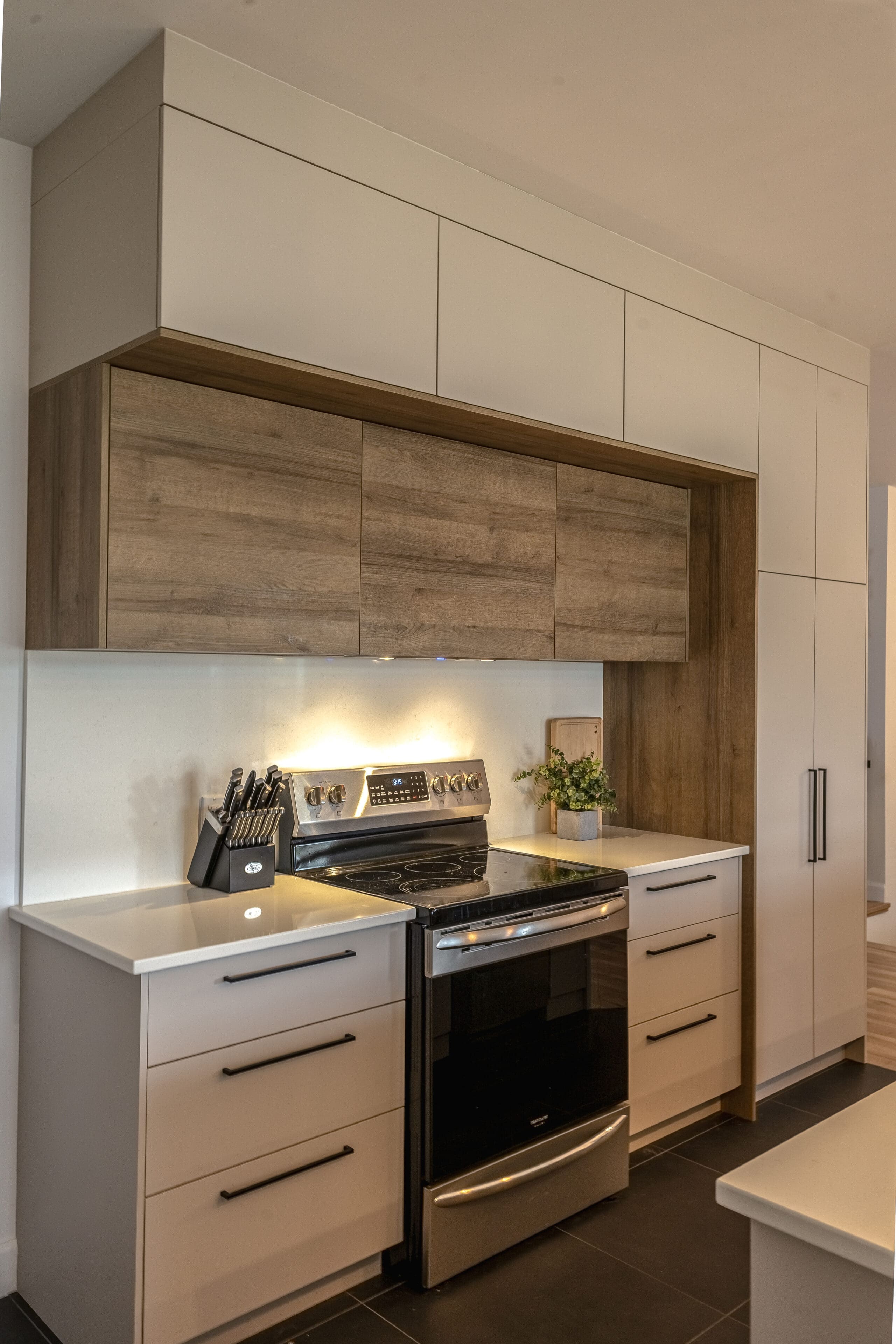 Cuisine moderne avec des armoires blanches, des poignées noires, des armoires supérieures en bois, une cuisinière électrique avec four, un bloc de couteaux et une petite plante sur le comptoir. L'espace est propre et bien éclairé.