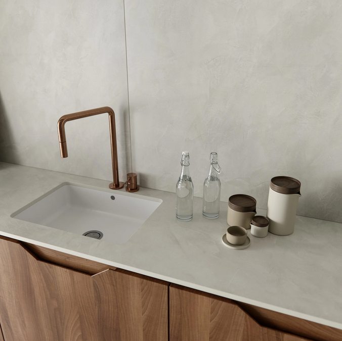 Un comptoir de cuisine moderne avec un évier carré, un robinet en bronze, deux bouteilles en verre avec bouchon, et plusieurs récipients beiges avec des couvercles bruns, sur un mur de couleur claire et des armoires en bois.