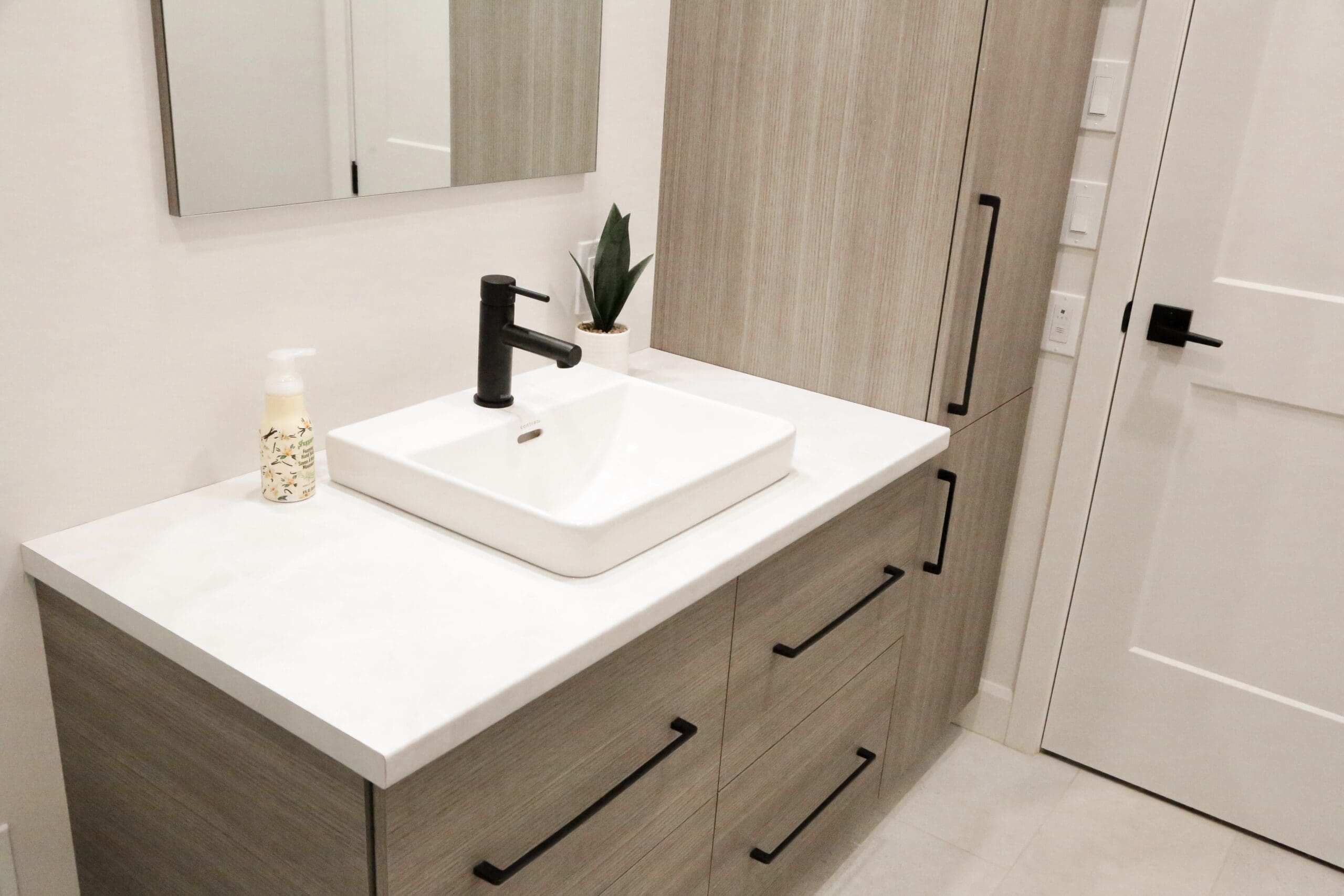 Meuble de salle de bain moderne avec un lavabo rectangulaire blanc, un robinet noir, un distributeur de savon, une petite plante, un grand miroir et des armoires en bois clair avec des poignées noires. La porte blanche et les murs clairs sont visibles.