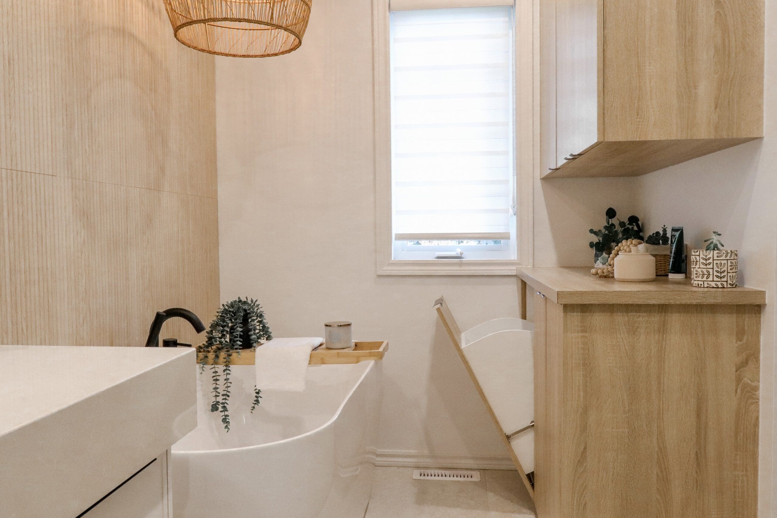 Salle de bains moderne avec une baignoire blanche autoportante, un plateau en bois, une serviette et une plante verte. Des armoires en bois clair, un panier à linge et une fenêtre avec un abat-jour blanc ajoutent au design épuré et minimaliste.