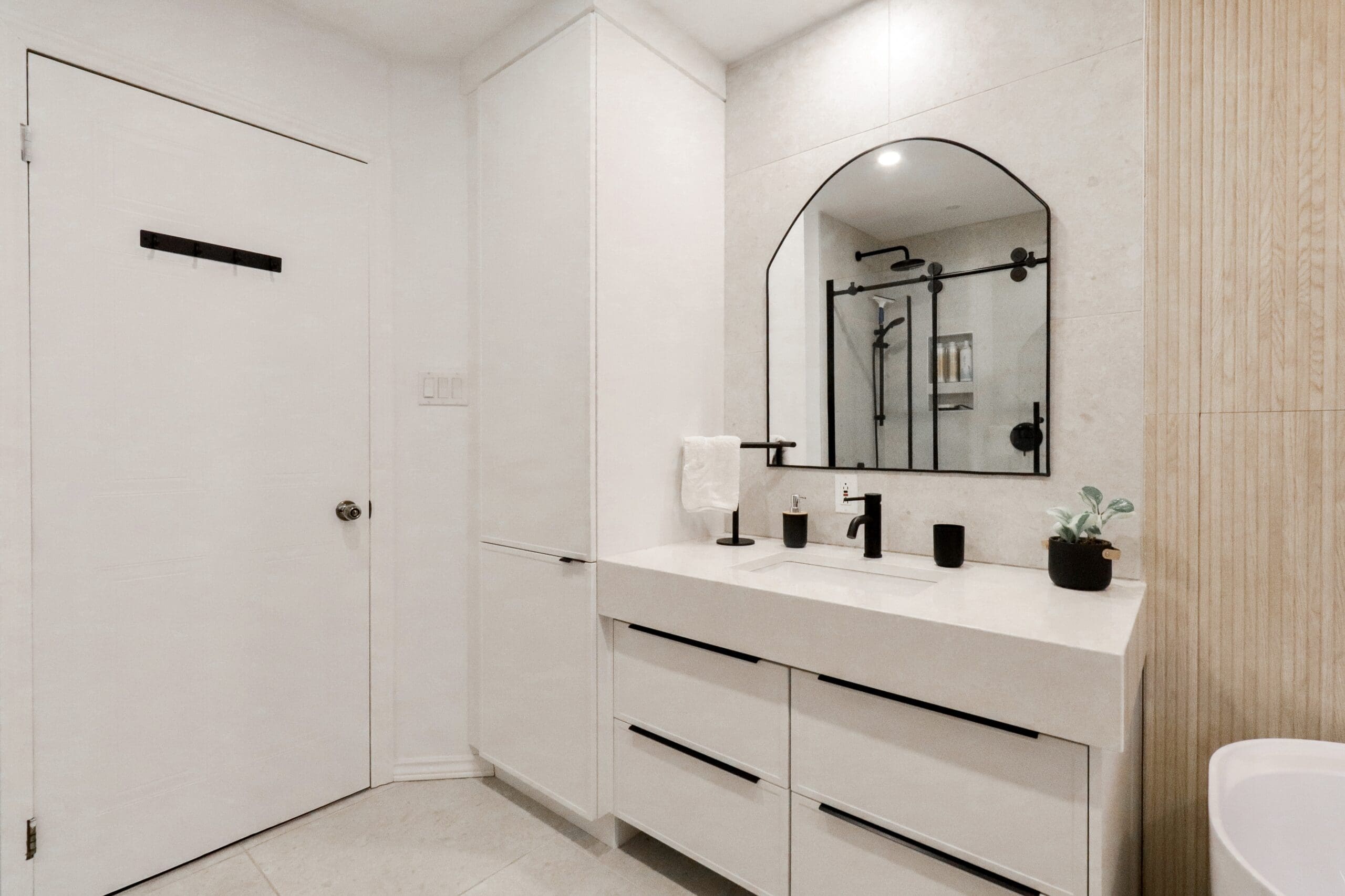Une salle de bains moderne avec des armoires blanches, un grand lavabo rectangulaire, un robinet noir, un miroir arqué encadré de noir et un décor minimal comprenant une petite plante et des distributeurs de savon. Une porte blanche est visible à gauche.