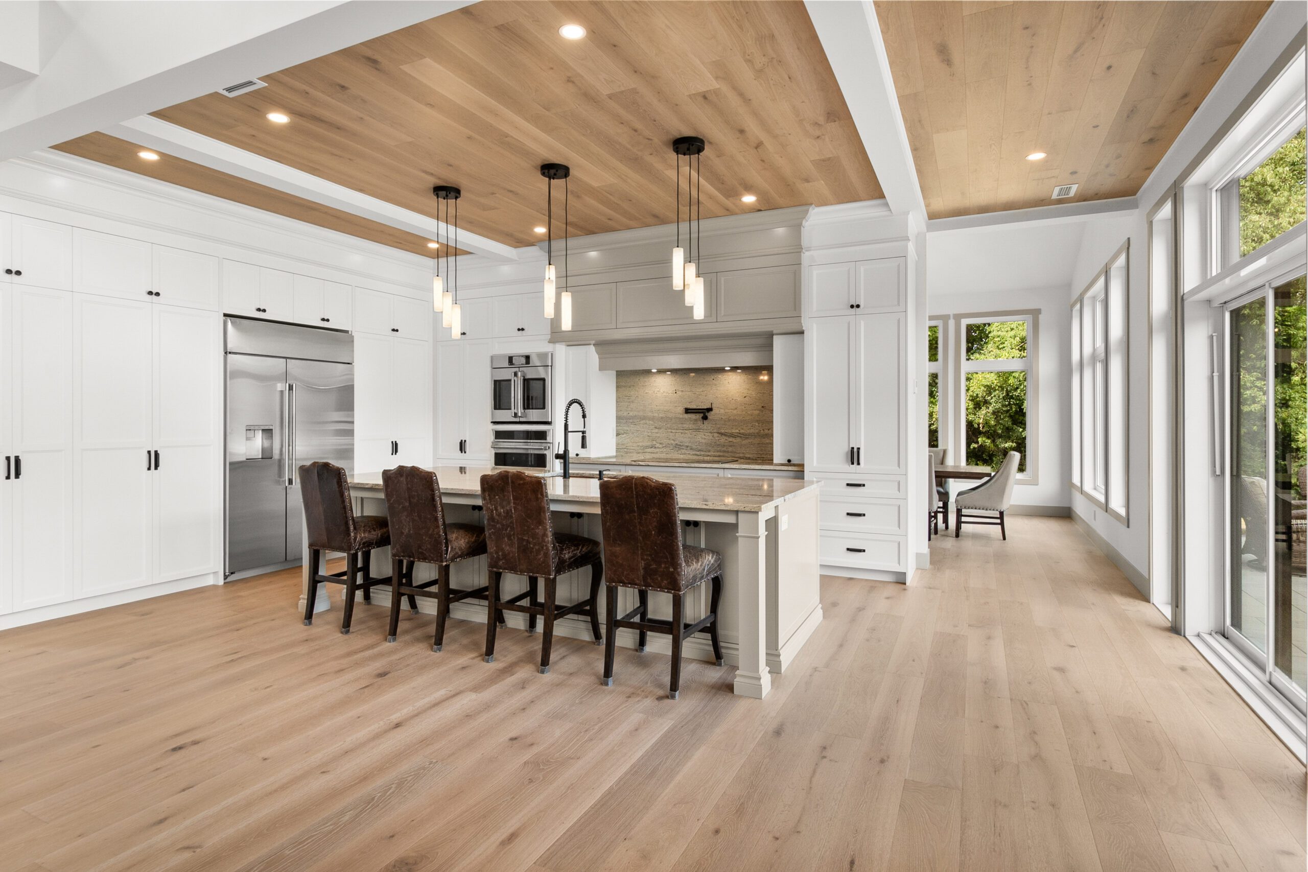 Cuisine moderne avec un parquet en bois clair, des placards blancs, des appareils électroménagers en inox, un grand îlot équipé de chaises, des suspensions et de grandes fenêtres laissant entrer la lumière naturelle.