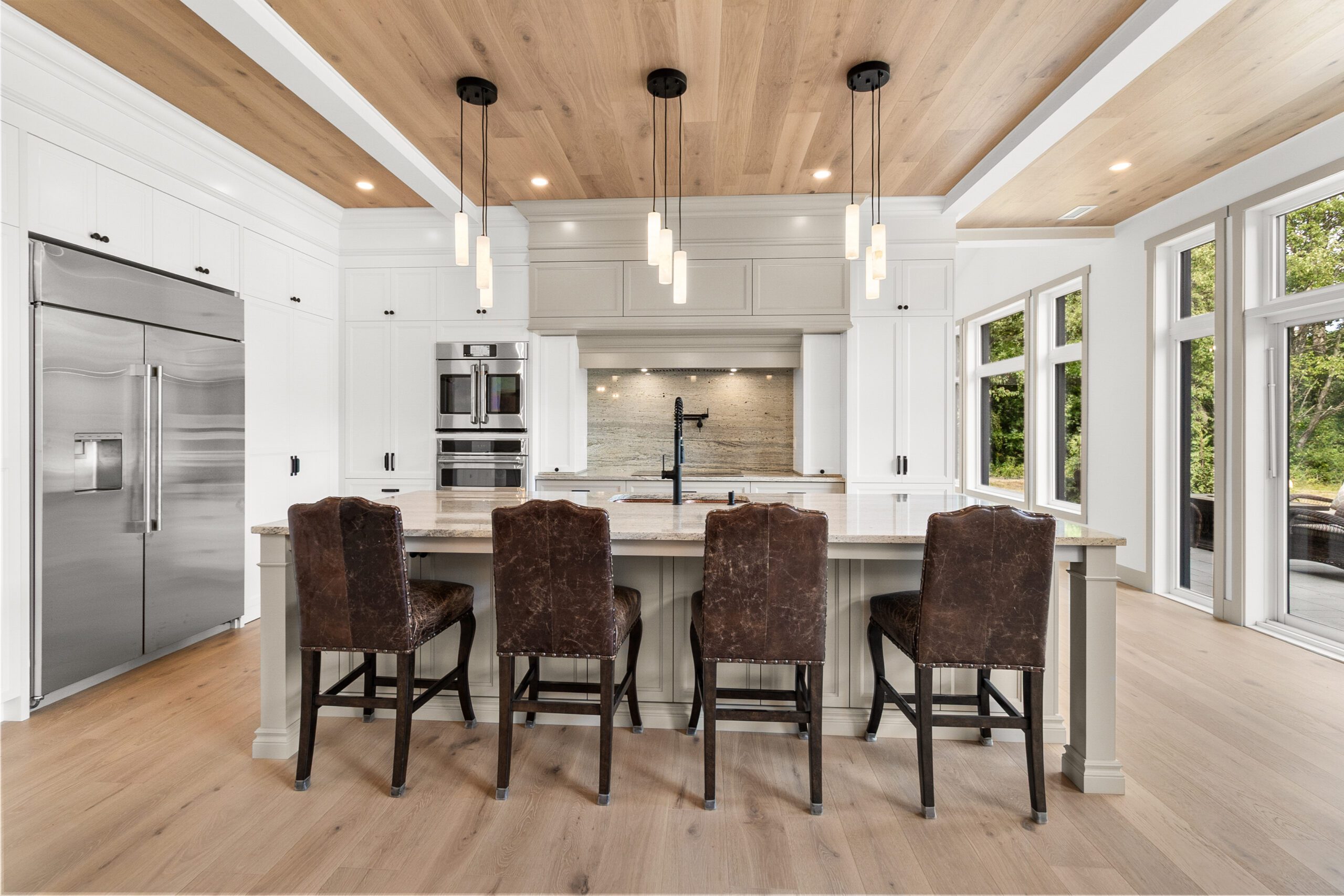 Cuisine moderne avec un grand îlot, quatre chaises en cuir foncé, des appareils électroménagers en inox, des placards blancs, des suspensions, un plafond en bois et de grandes fenêtres donnant sur l'extérieur.