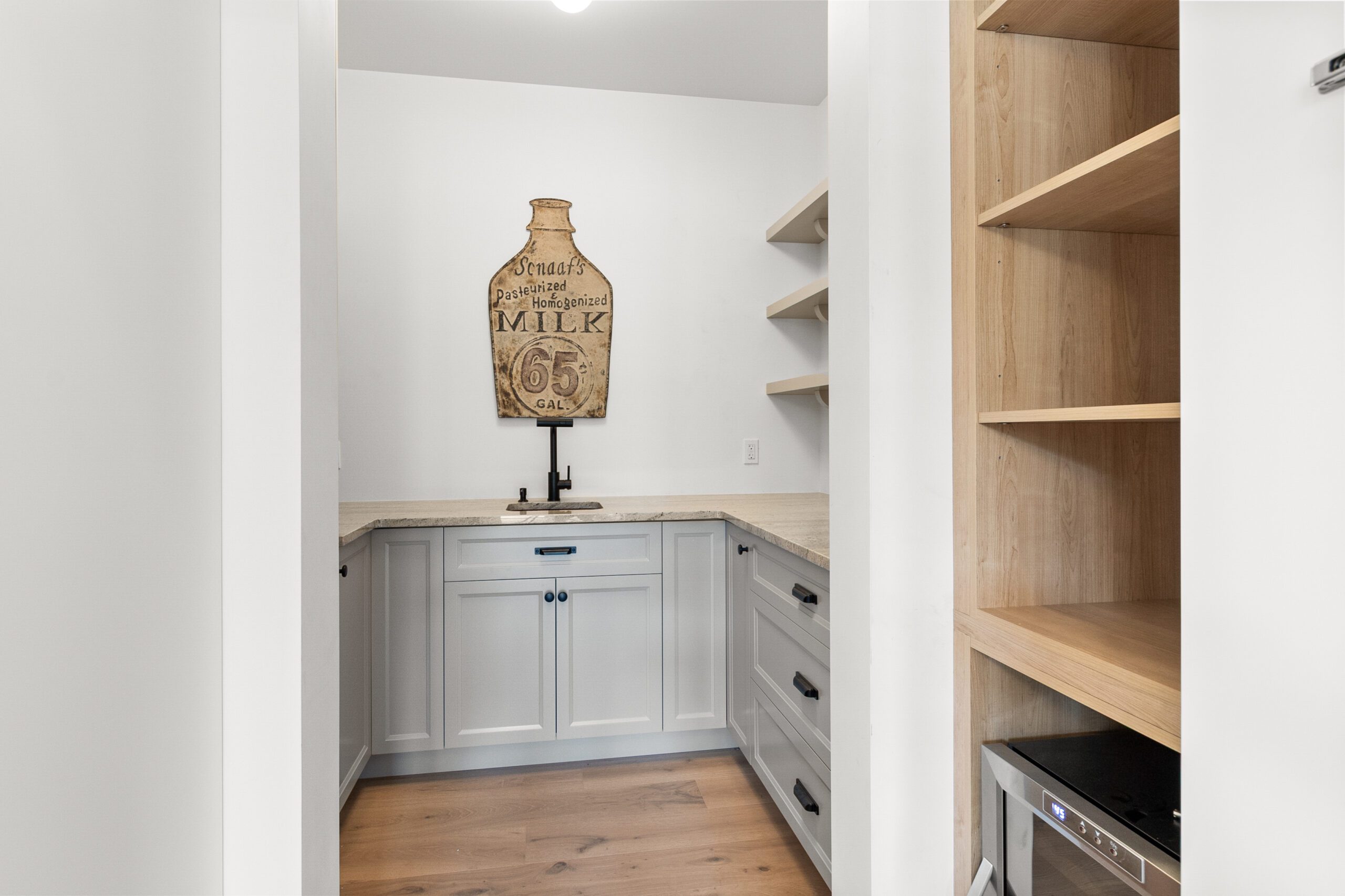 Un petit cellier avec des placards gris clair, un robinet noir, des étagères ouvertes, du parquet et une enseigne vintage représentant un verre de lait accrochée au mur au-dessus de l'évier. On aperçoit un appareil électroménager et des étagères en bois sur la droite.