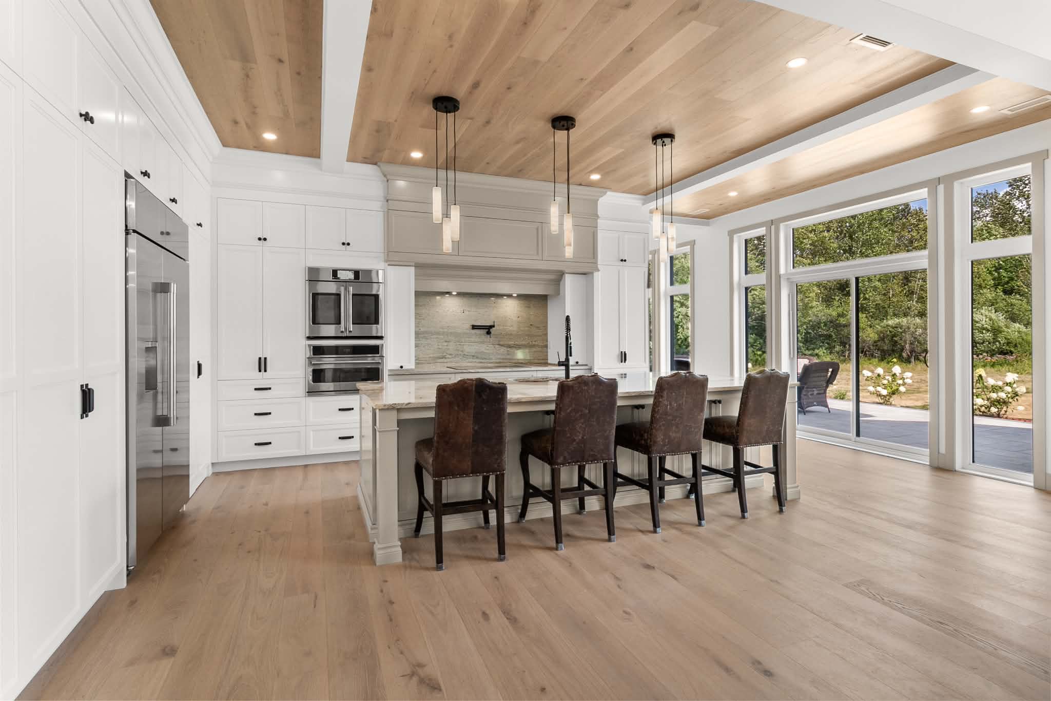 Cuisine moderne avec des placards blancs, des appareils électroménagers en inox, un plafond en bois, un grand îlot avec quatre chaises foncées, des suspensions et des baies vitrées donnant sur un jardin verdoyant.